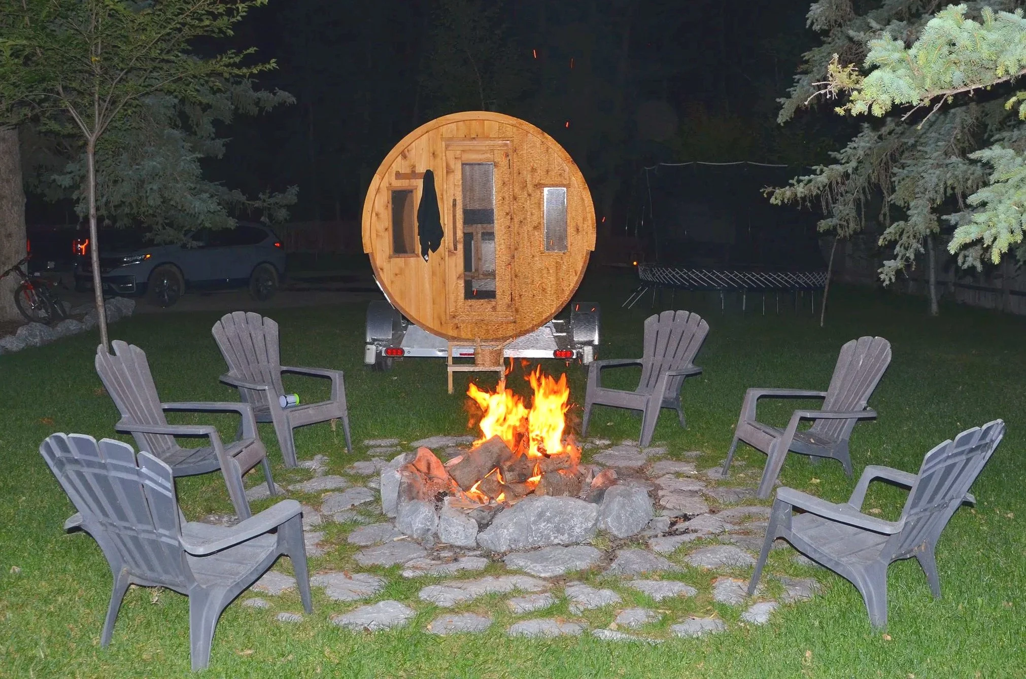Nighttime backyard scene with a fire pit surrounded by chairs, and a wooden barrel sauna