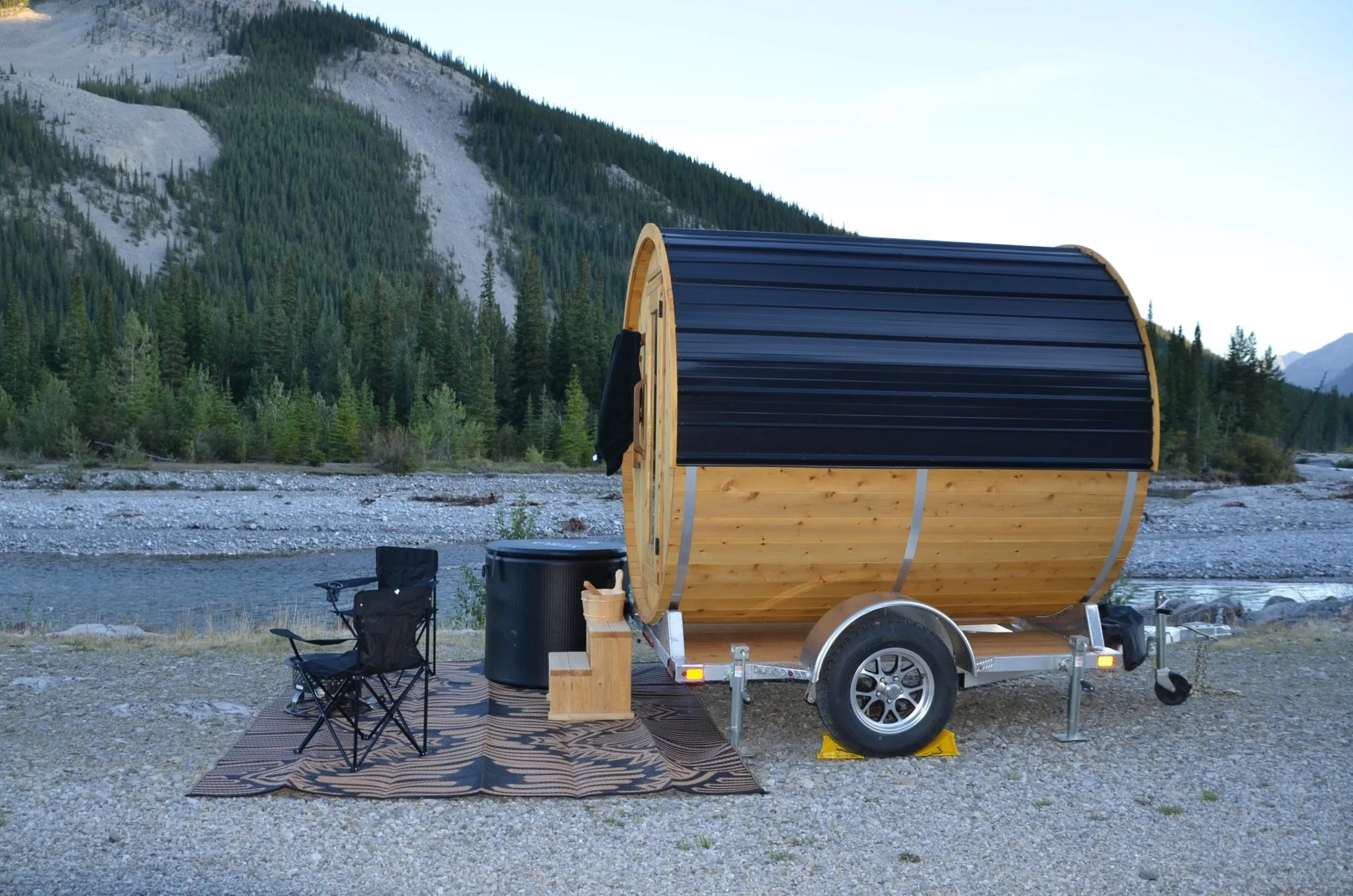 A wooden barrel sauna with a black curved roof and wooden sides parked on a gravel area by a river. There are folding chairs, a small rug, a black cold plunge tub, and outdoor gear nearby, with a backdrop of forested mountains and a clear sky.