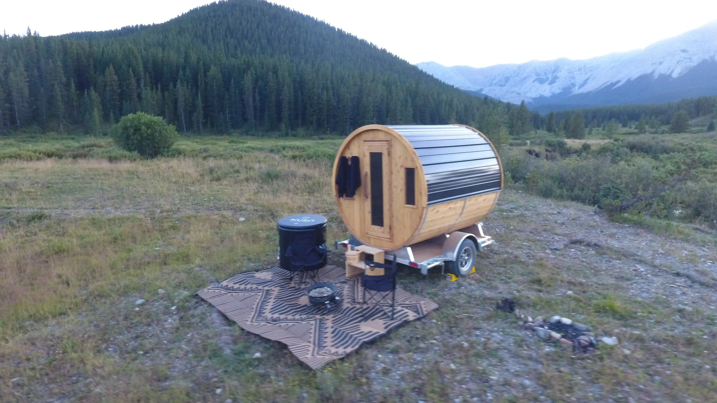 A wooden barrel-shaped sauna on a trailer, set in an open field with mountains and pine trees in the background. There is a black cold plunge tub, a patterned outdoor rug, a camping chair, and a fire pit nearby.