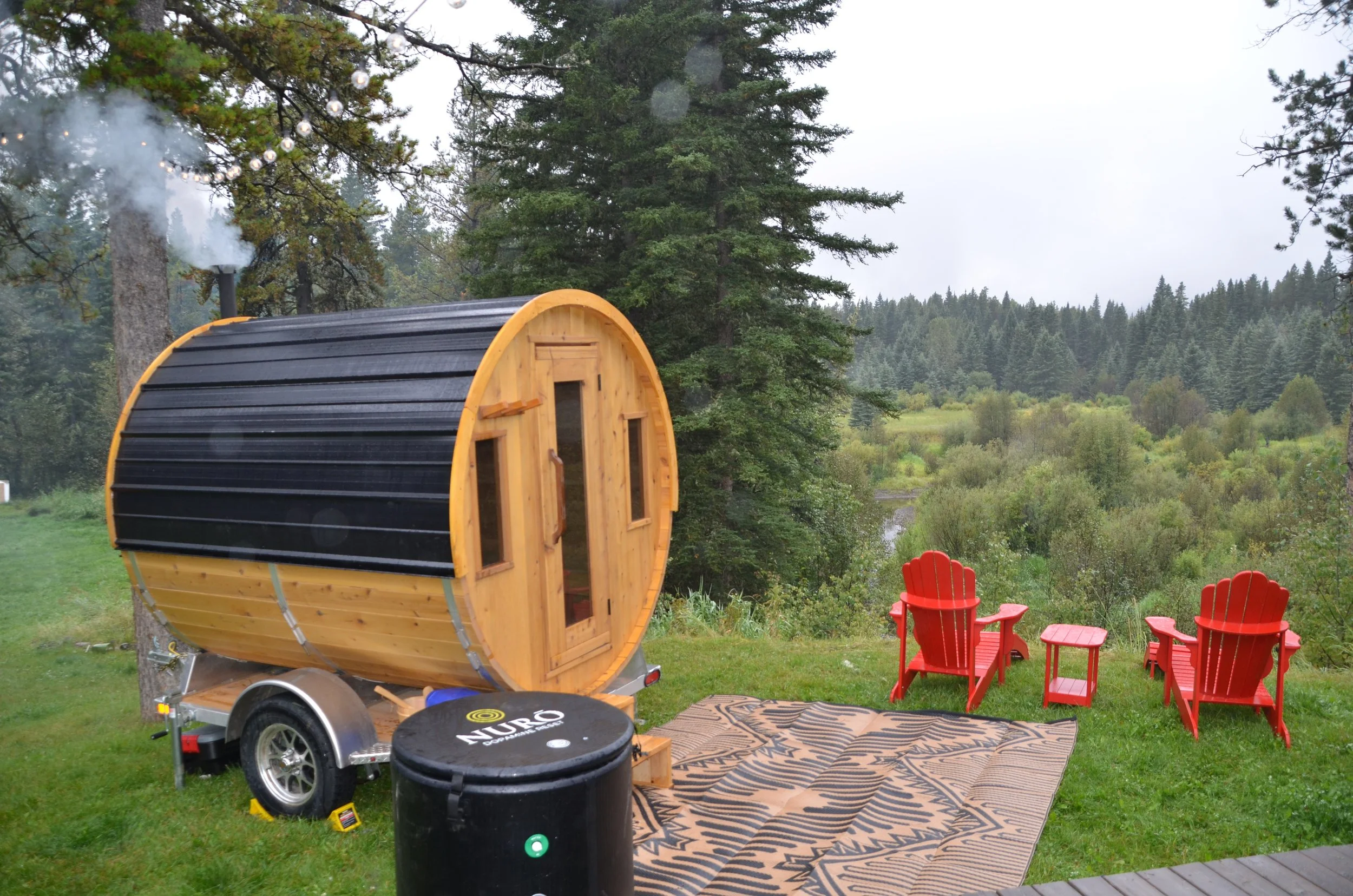 A wooden sauna with a curved black roof is parked outdoors among trees. To the right, there are three red Adirondack chairs on a patterned outdoor rug. In the background, there is a scenic view of a forested area with tall trees, and a misty sky.