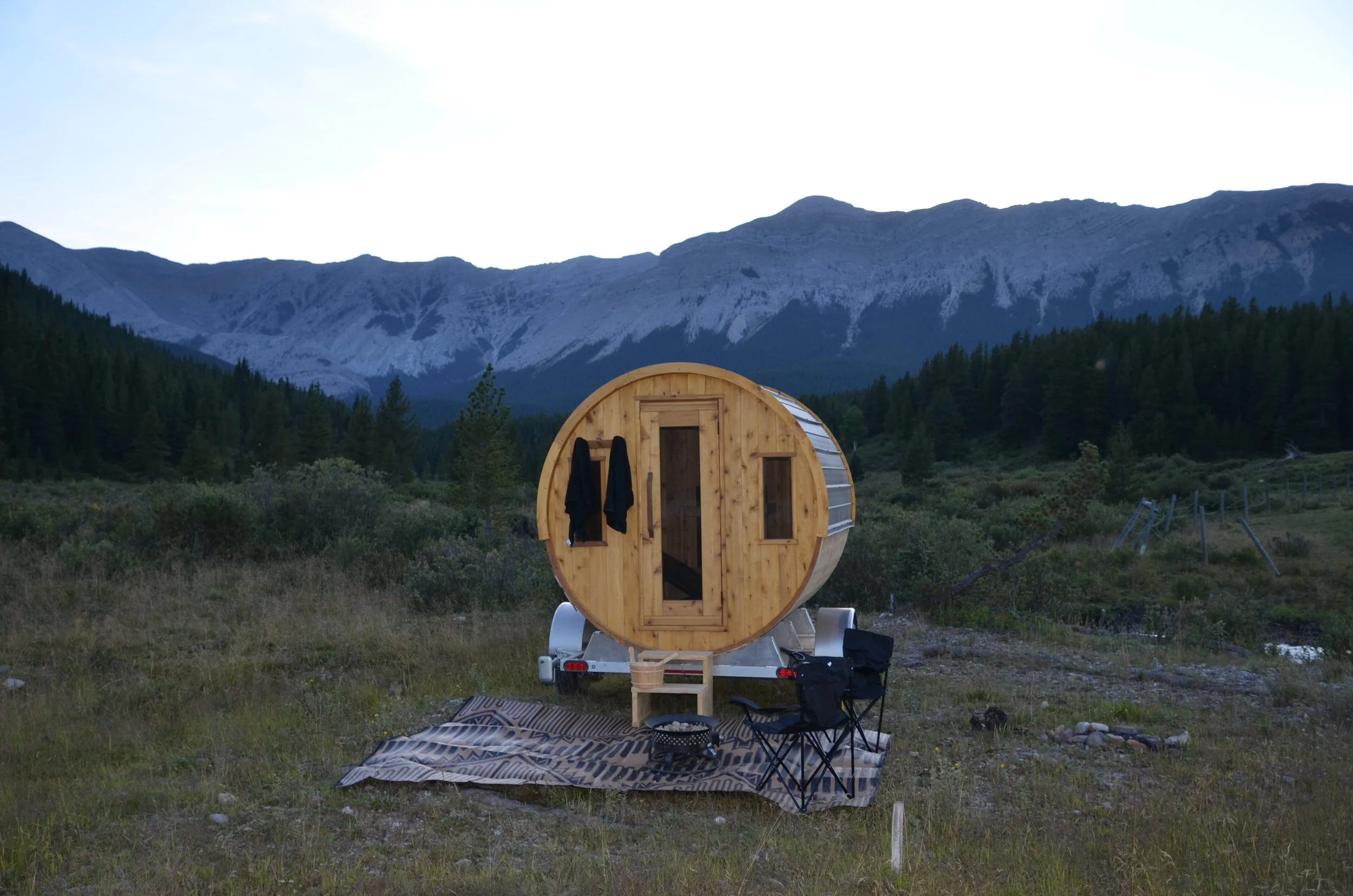 A wooden barrel sauna parked on grass with a mountain range in the background. Two black towels hang on the side of the door, and camping chairs are set in front of it on a patterned mat.