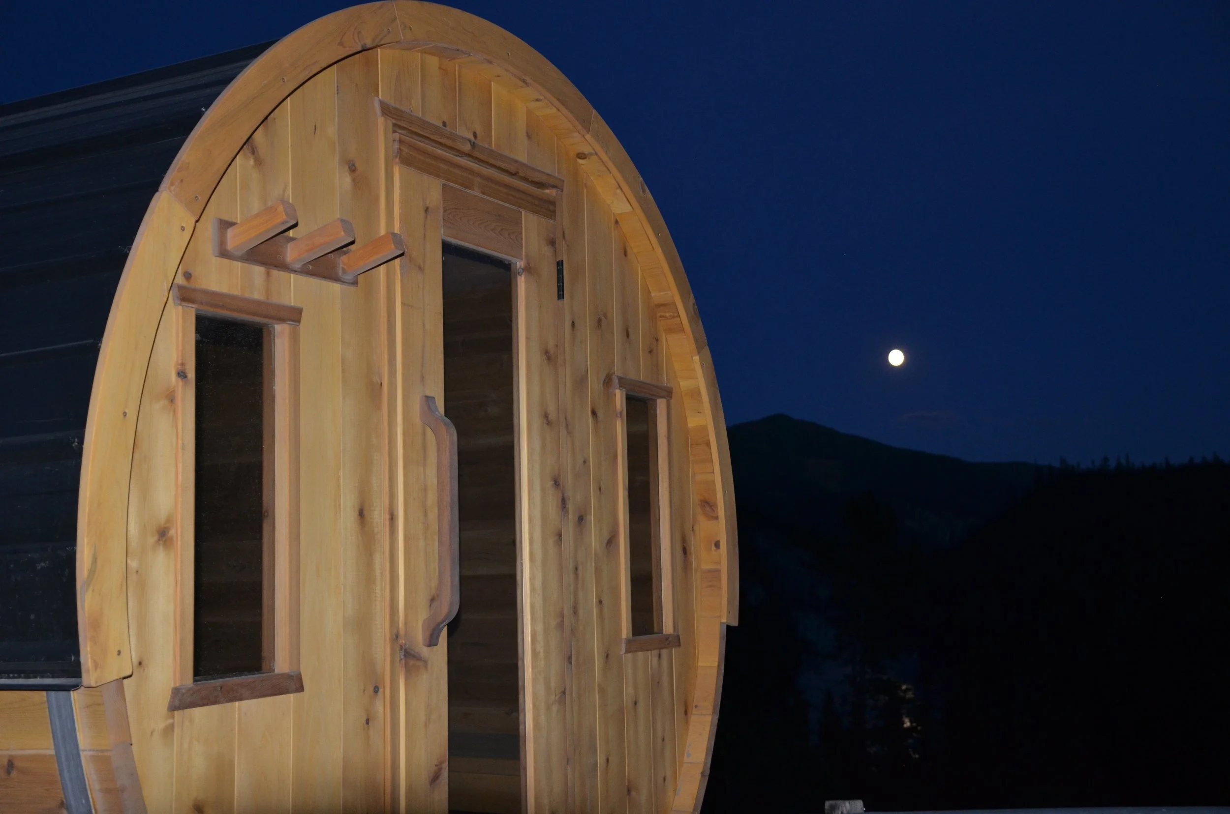 Night sky with a bright moon over a wooden barrel sauna with small door and two windows on either side.