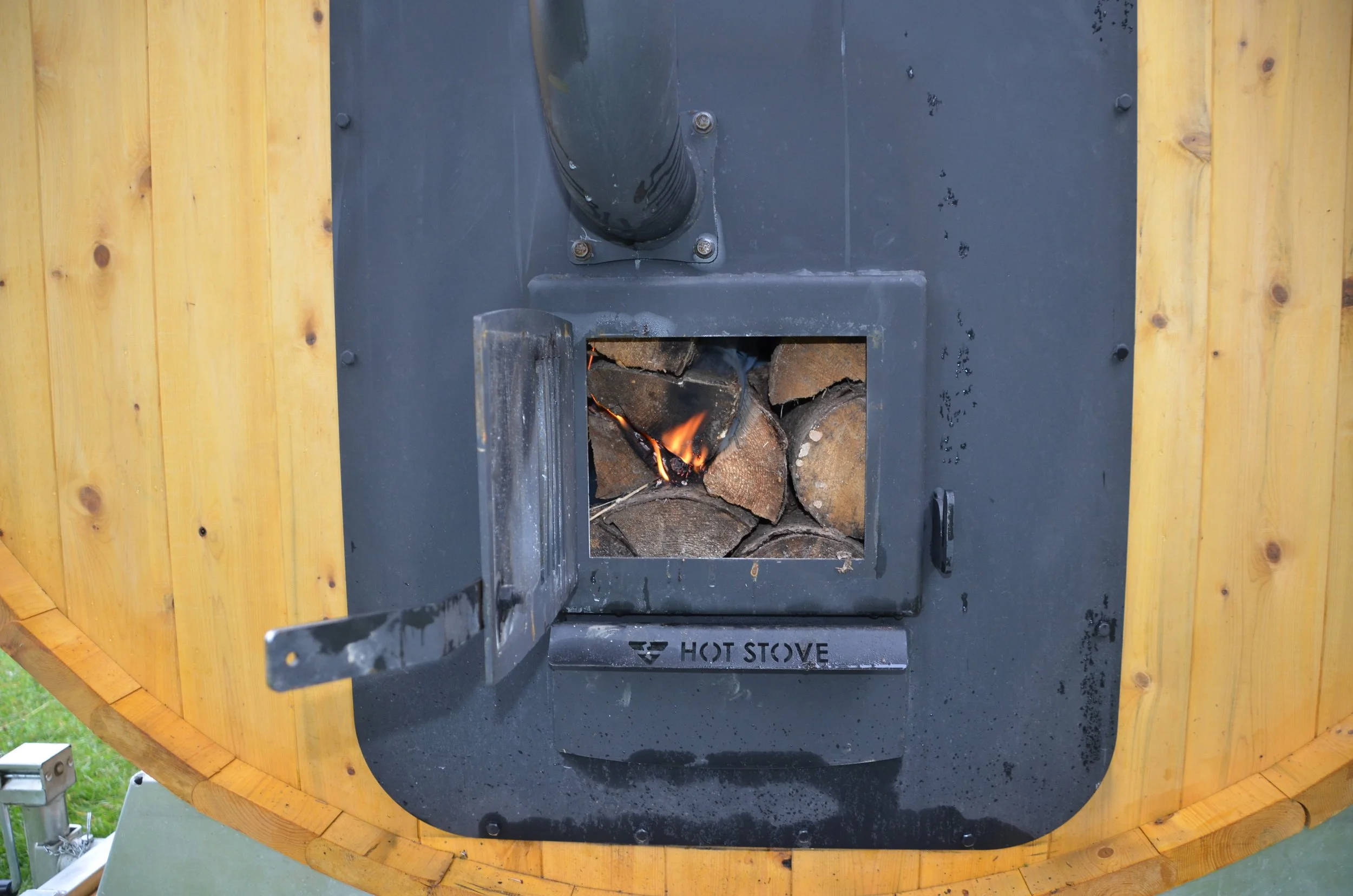 A small black metal wood stove with an open door showing burning logs inside, mounted in the back of a round barrel sauna.