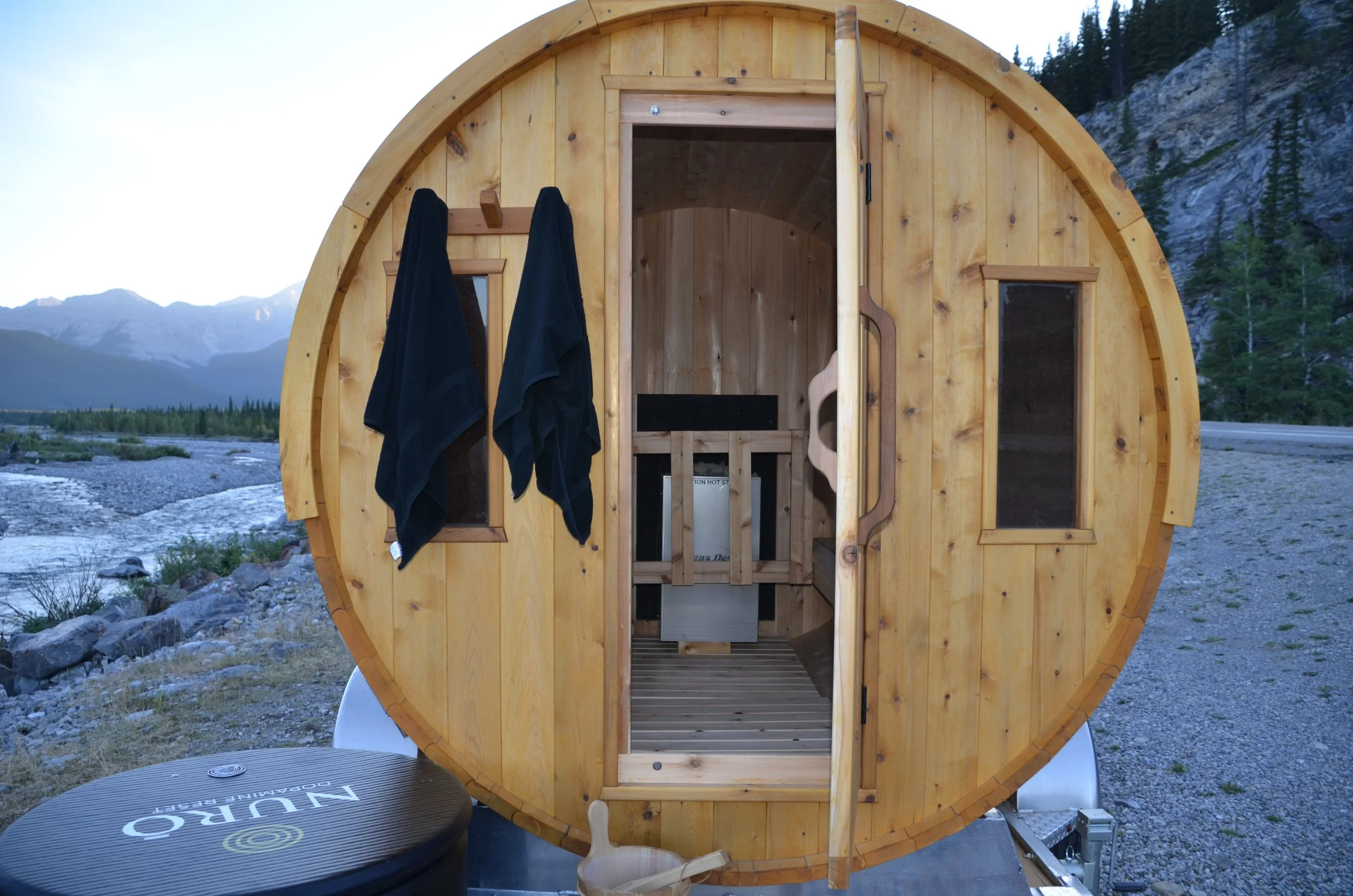 A wooden barrel shaped sauna sits outdoors near a river and mountains in the background. The open door reveals an interior with wooden walls and a small bench. Two black towels hang on a wooden rack outside the sauna.