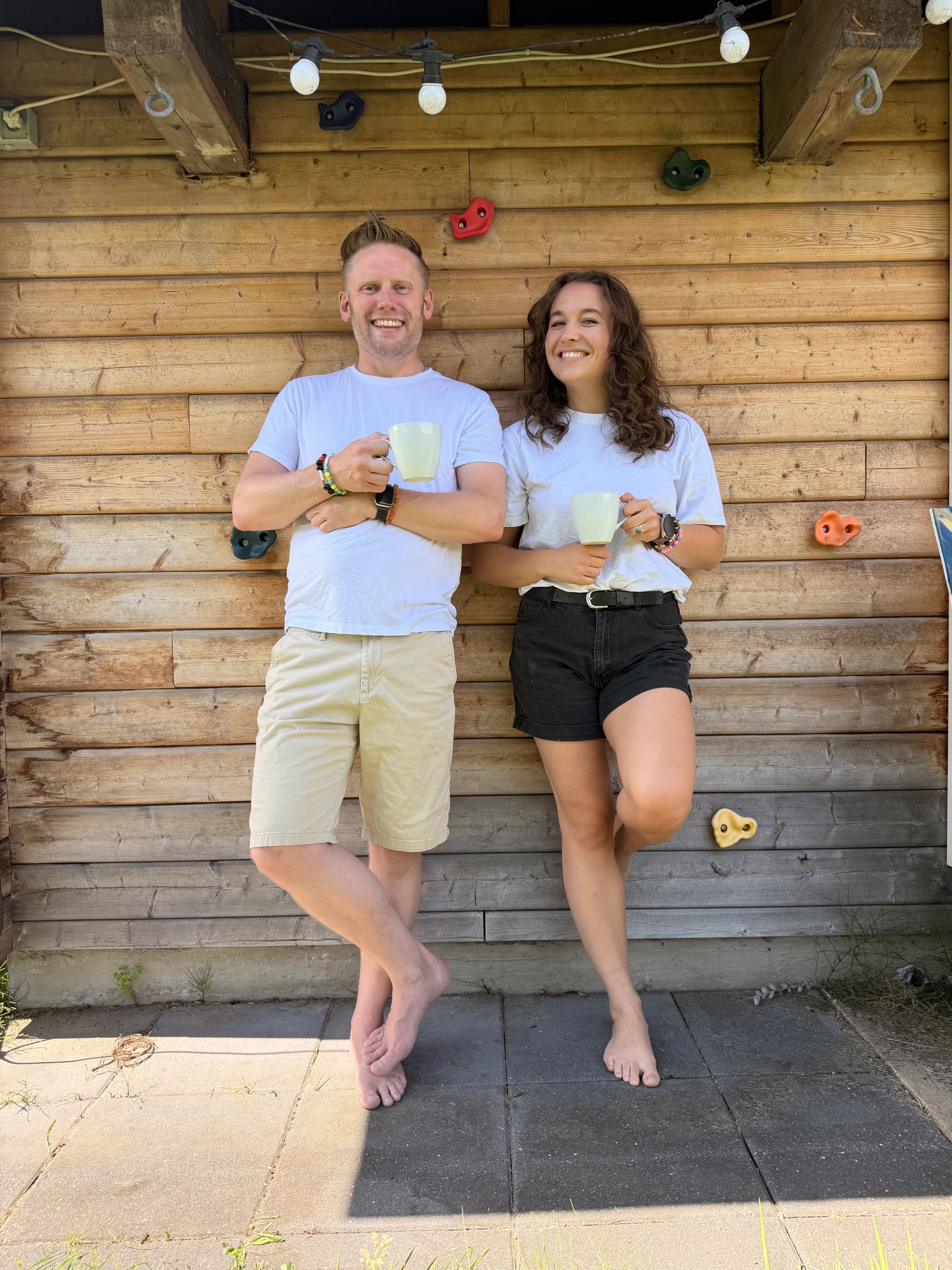 Two smiling people standing barefoot against a wooden wall with colorful climbing holds. The person on the left is a male with short hair, a beard, wearing a white T-shirt, beige shorts, and has beaded bracelets. The person on the right is a female with curly hair, wearing a white T-shirt, black shorts, and a watch. They are holding white mugs.