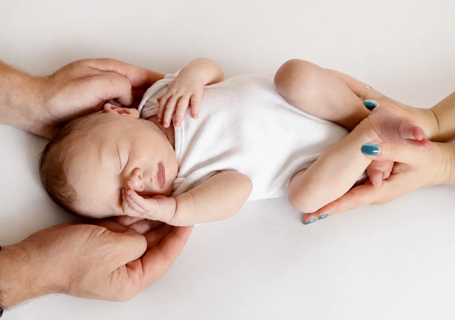 A newborn baby sleeping on a white surface, gently held and supported by two adults' hands.