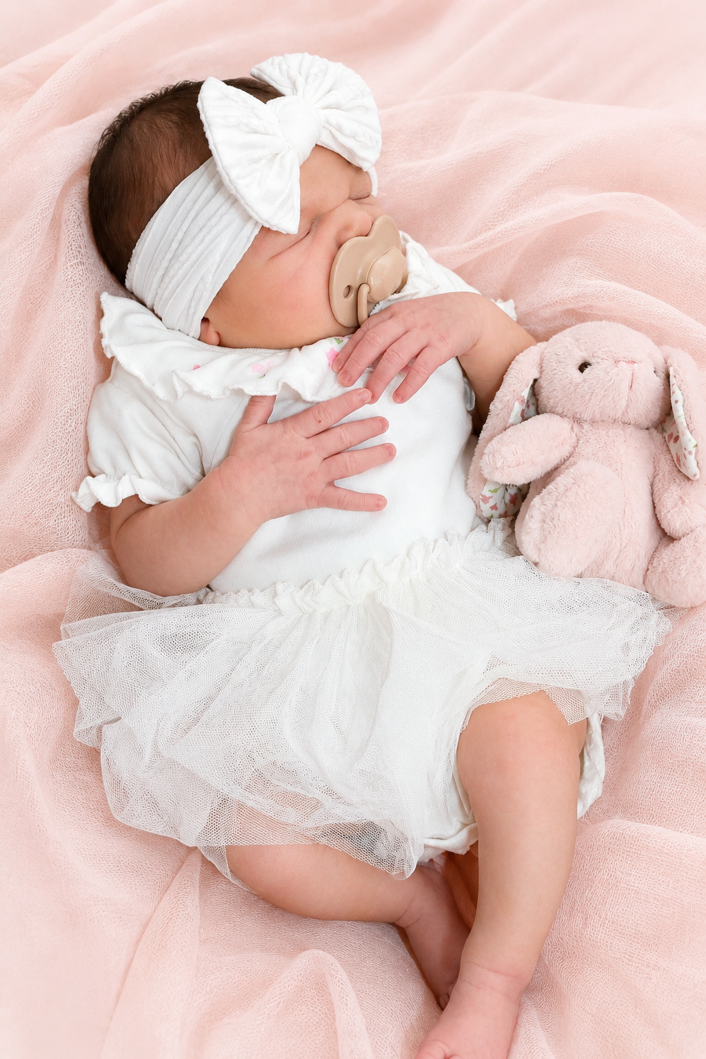 A sleeping baby girl with a pacifier, wearing a headband with a bow, lying on pink fabric next to a pink stuffed bunny, dressed in a white outfit with ruffled sleeves and a tulle skirt.