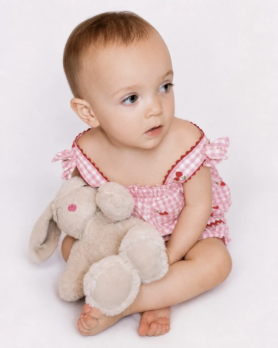 A young girl sitting with an off-white stuffed bunny, wearing a pink checkered dress, looking to the side with a neutral expression.
