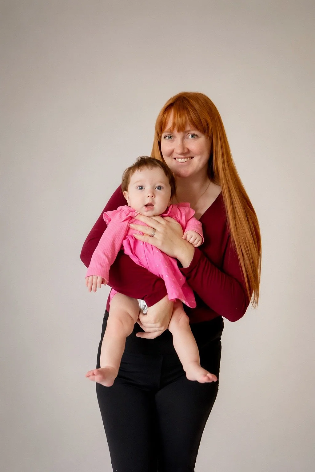A woman with long red hair holding a baby girl with brown hair; both are smiling, with the baby wearing a pink dress and the woman wearing a maroon top, against a plain light-colored background.