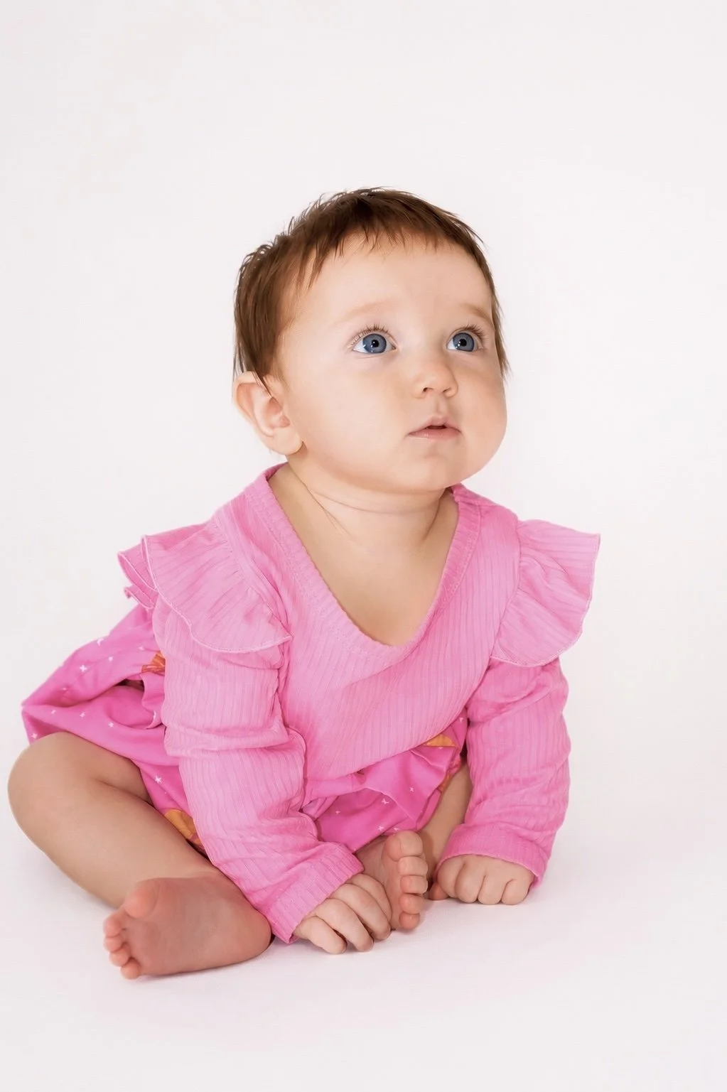 A baby girl with blue eyes and brown hair, wearing a pink dress and sitting on the floor with a white background.
