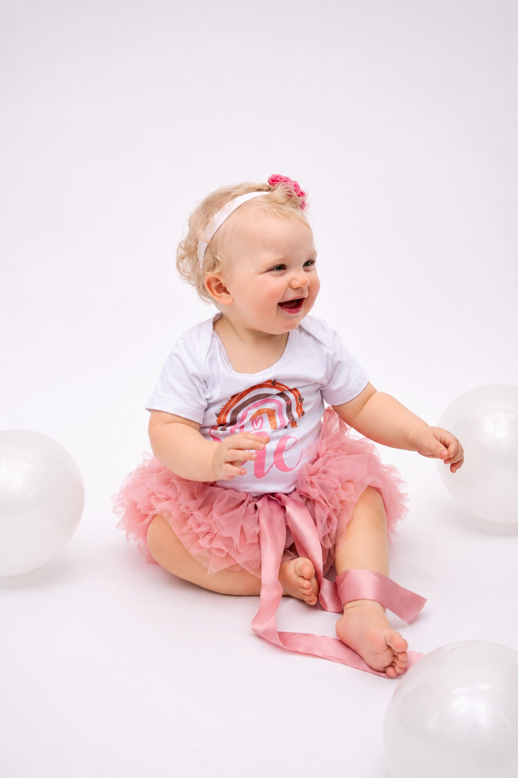 Young girl sitting on the floor wearing a white t-shirt with a rainbow and the word "love", a pink tutu, and a pink headband with a flower, surrounded by white balloons, smiling.