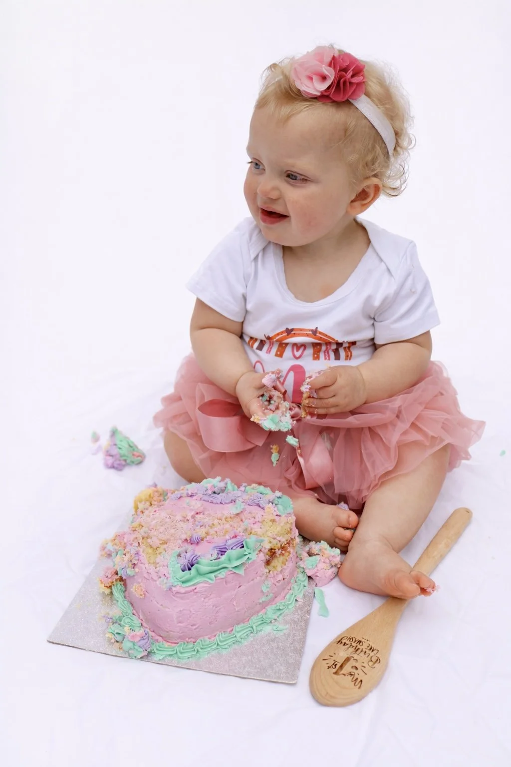 A young girl with blonde curly hair, wearing a pink flower headband, a white t-shirt, and a pink tutu, sitting on a white surface with a partially eaten pink and purple birthday cake in front of her.