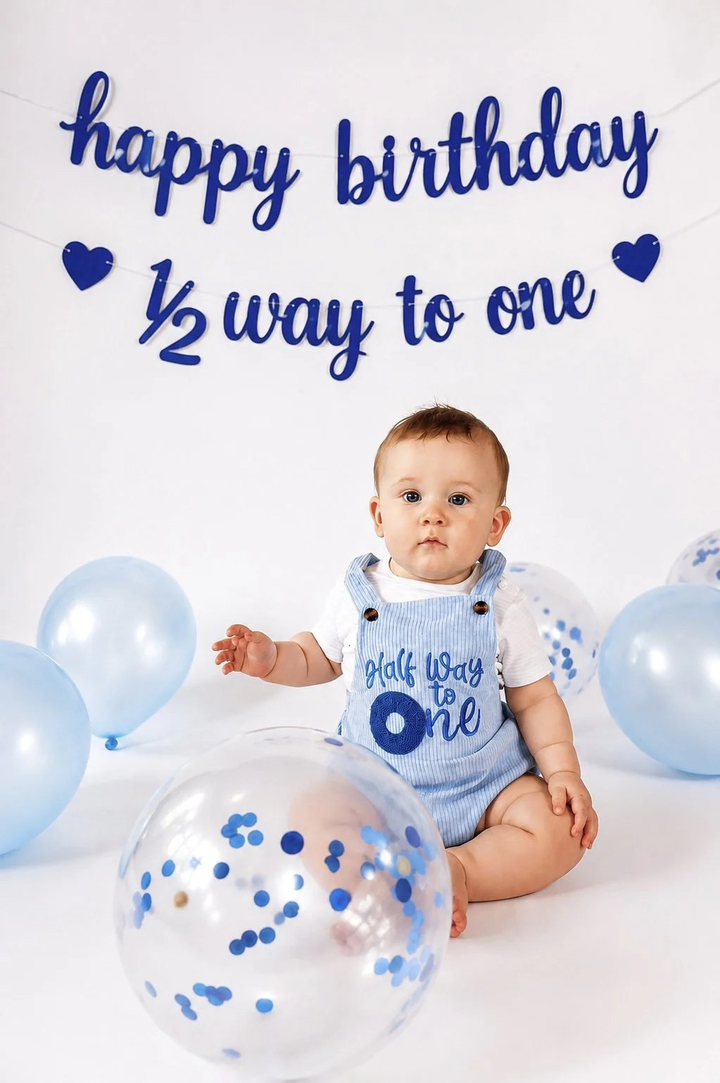 A baby sitting on the floor surrounded by blue balloons, with a white background and blue text that says "happy birthday ½ way to one." The baby is wearing a light blue romper with blue embroidered text that says "Half Way to One."