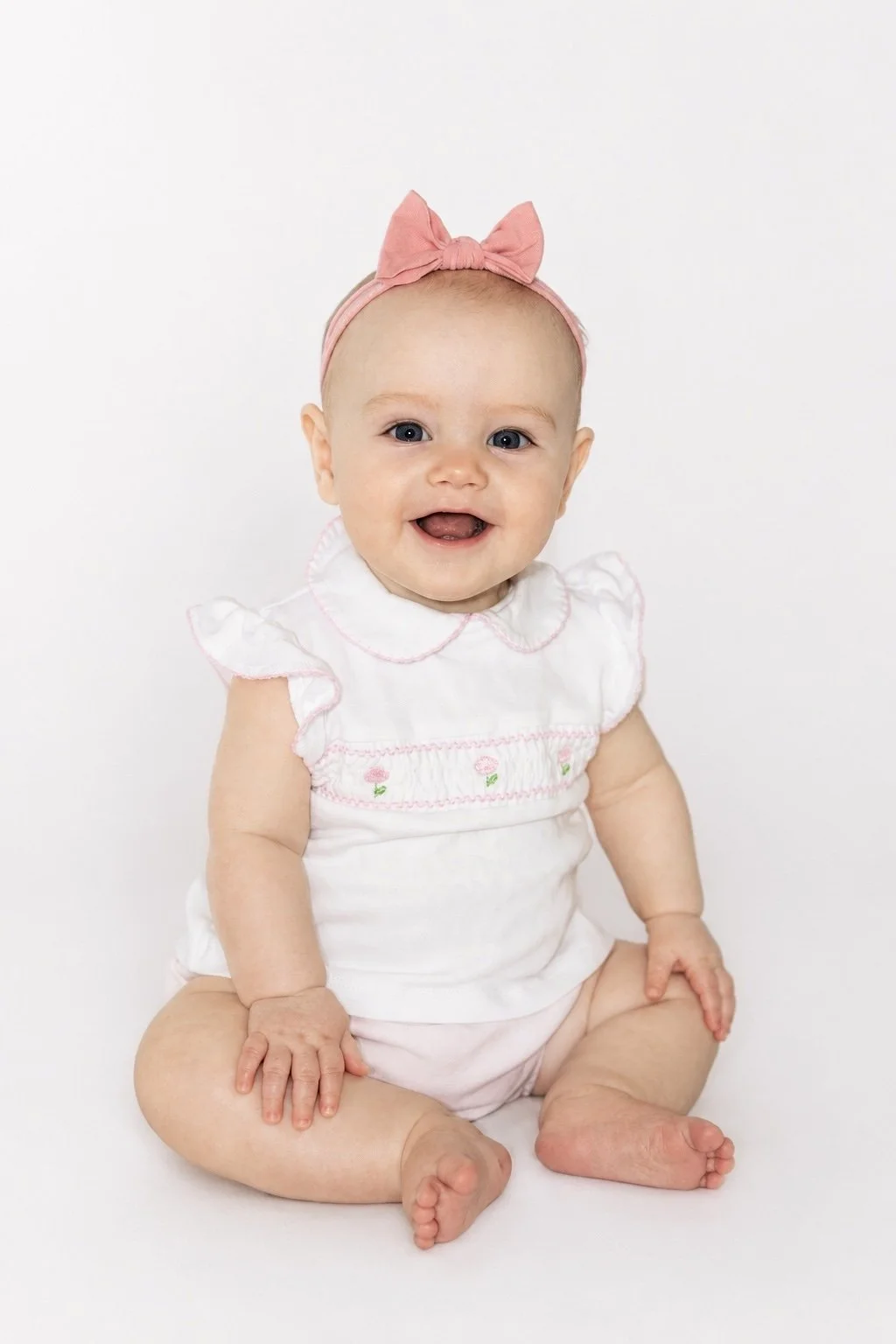 A smiling baby girl sitting on the floor against a white background, wearing a white dress with pink embroidery, a pink headband with a bow, and no shoes.