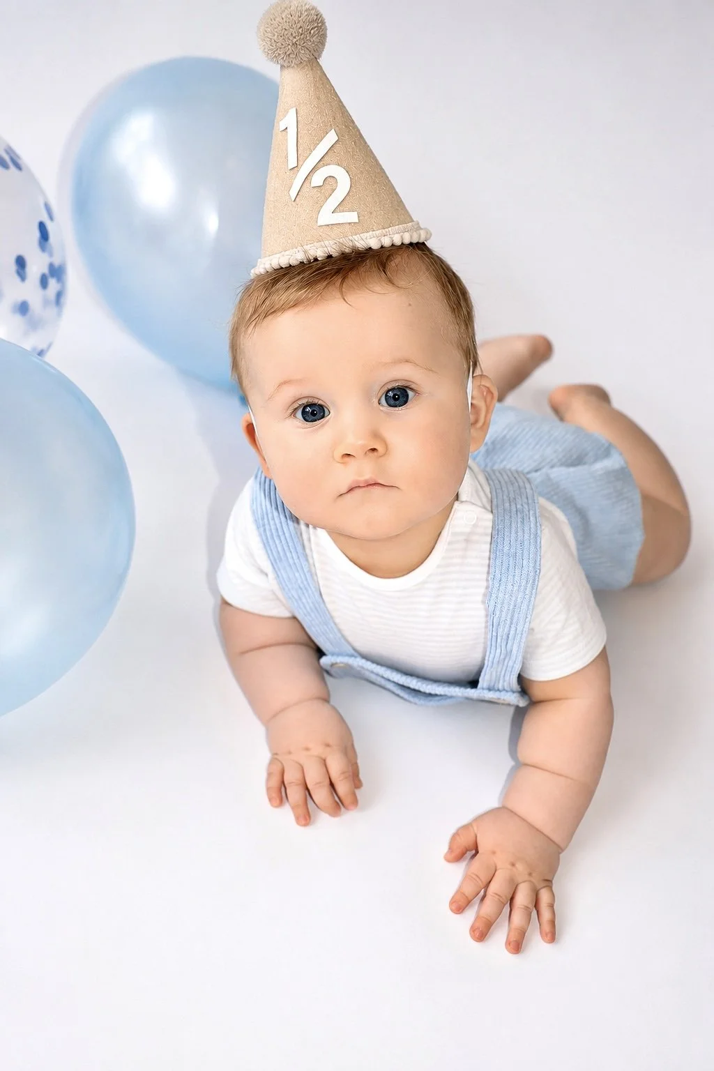 Baby laying on stomach wearing a birthday hat with '1/2' written on it, surrounded by blue balloons.