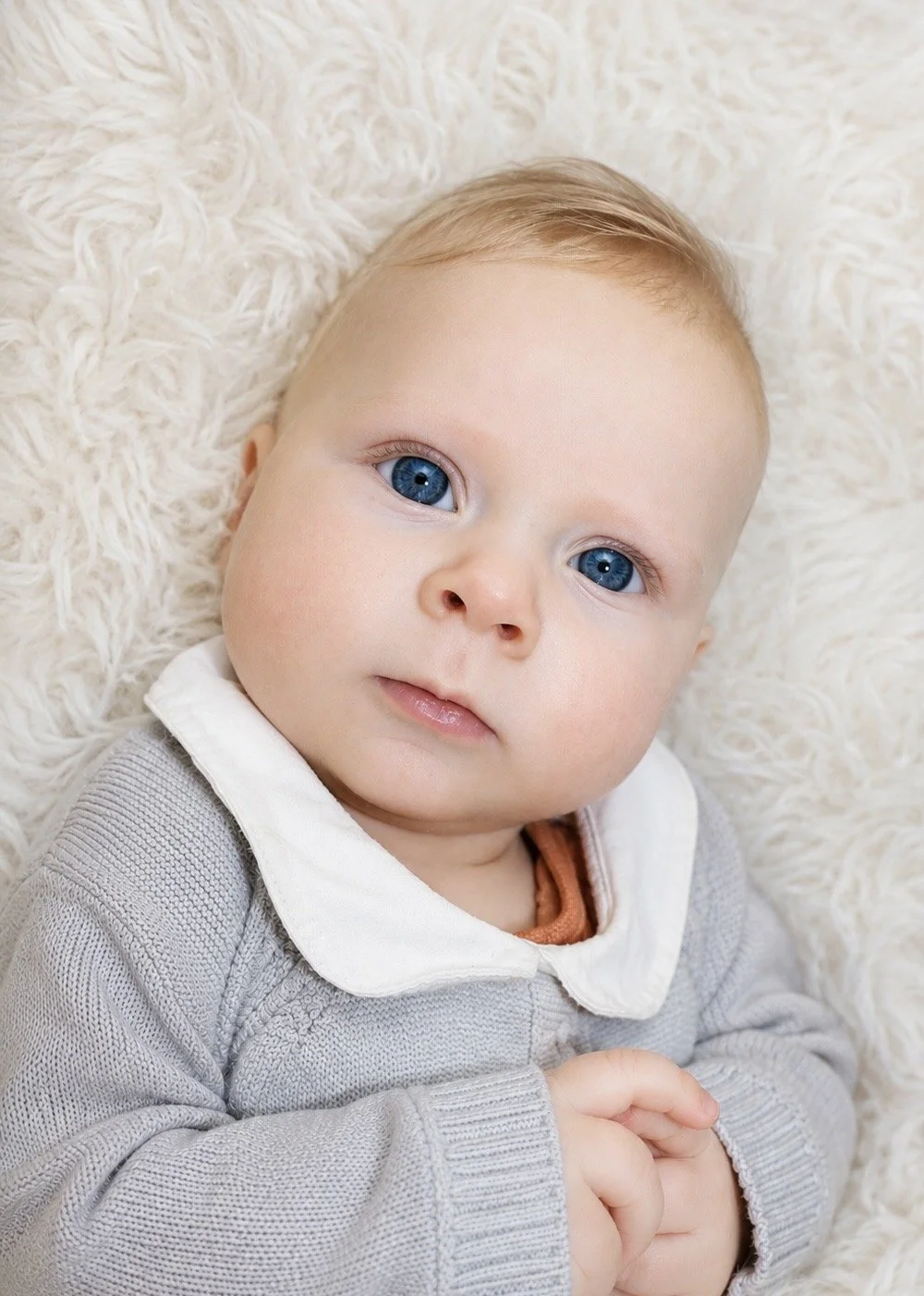 Close-up of a young boy with blue eyes, light blonde hair, wearing a gray sweater with a white collar, lying on a soft, cream-colored fluffy blanket.