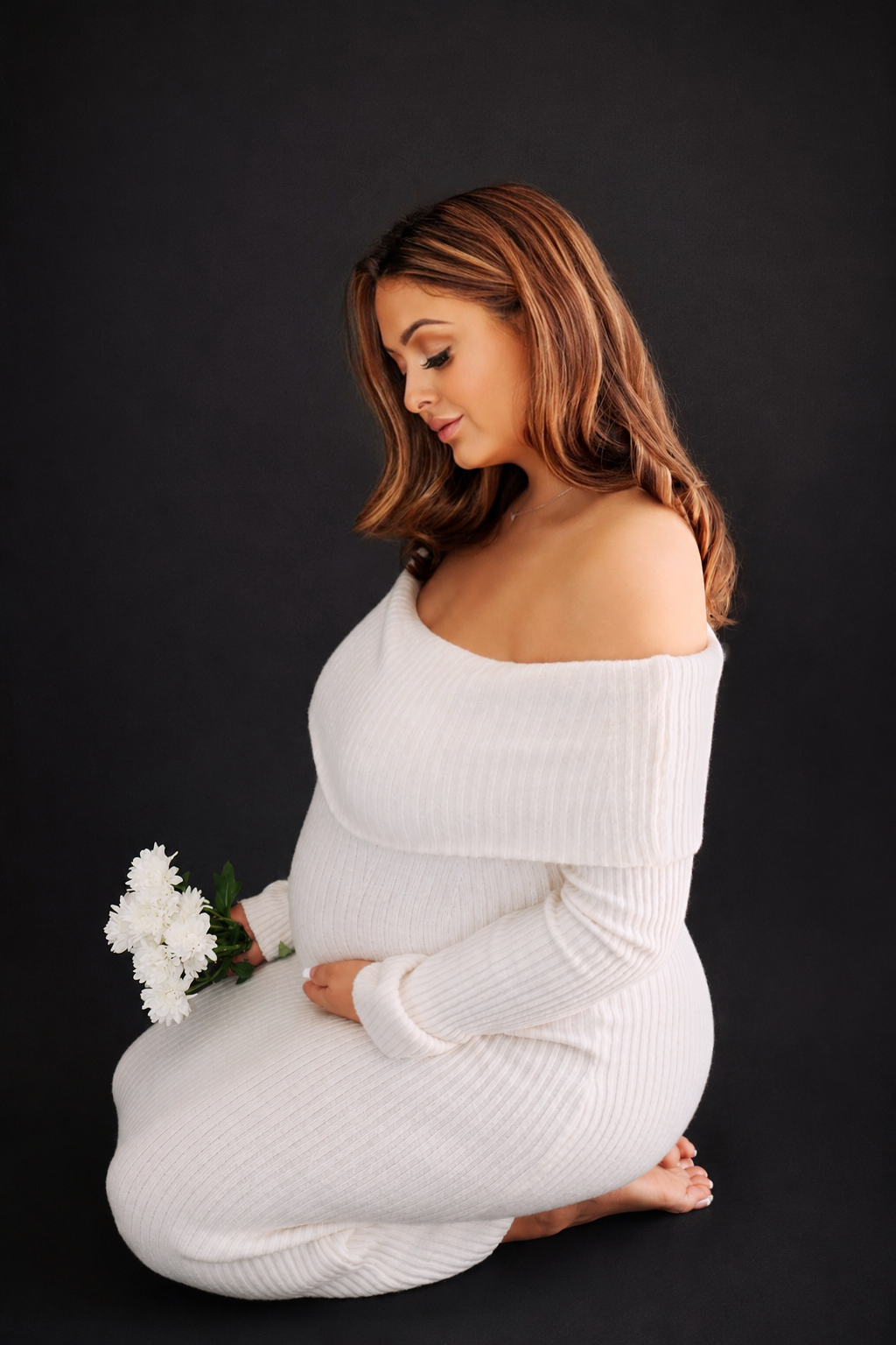 Pregnant woman with shoulder-length brown hair wearing a white off-shoulder sweater, kneeling against a black background, holding a bouquet of white flowers, looking down with a gentle expression.