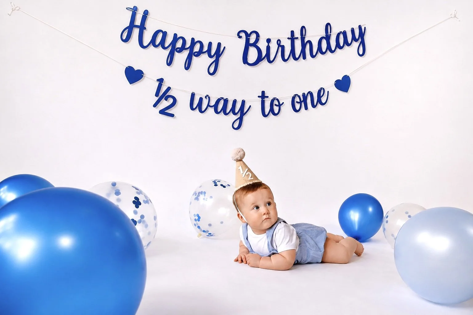 Baby lying on the floor in a birthday party setting with blue and white balloons, wearing a birthday hat with '1/2' on it, beneath a blue 'Happy Birthday 1/2 way to one' banner.