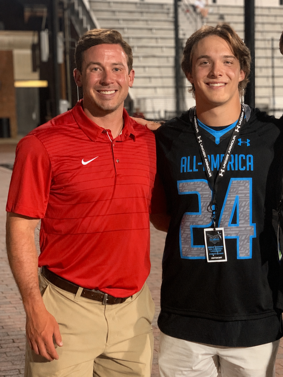Two men smiling, standing close together, one wearing a red Nike polo shirt and khaki pants, and the other wearing a black sports jersey with blue accents and a medal around his neck.