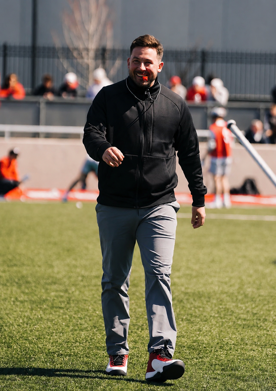 A man in a black athletic jacket and gray pants smiling while walking on a sports field, with several people in the background.