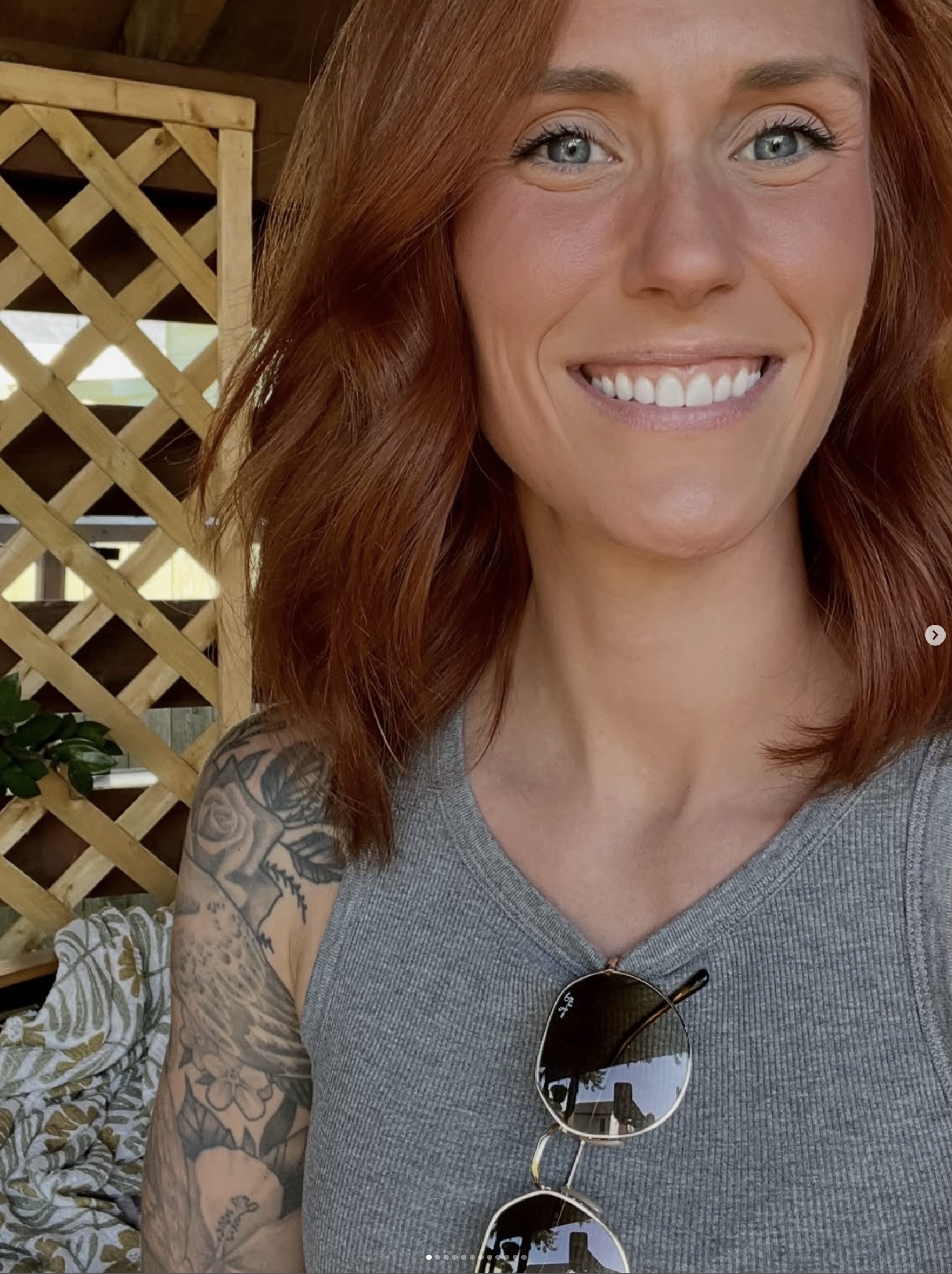 Close-up of a woman with red hair, blue eyes, and tattoos on her arm, smiling and wearing sunglasses on her chest, sitting outdoors near a wooden lattice.
