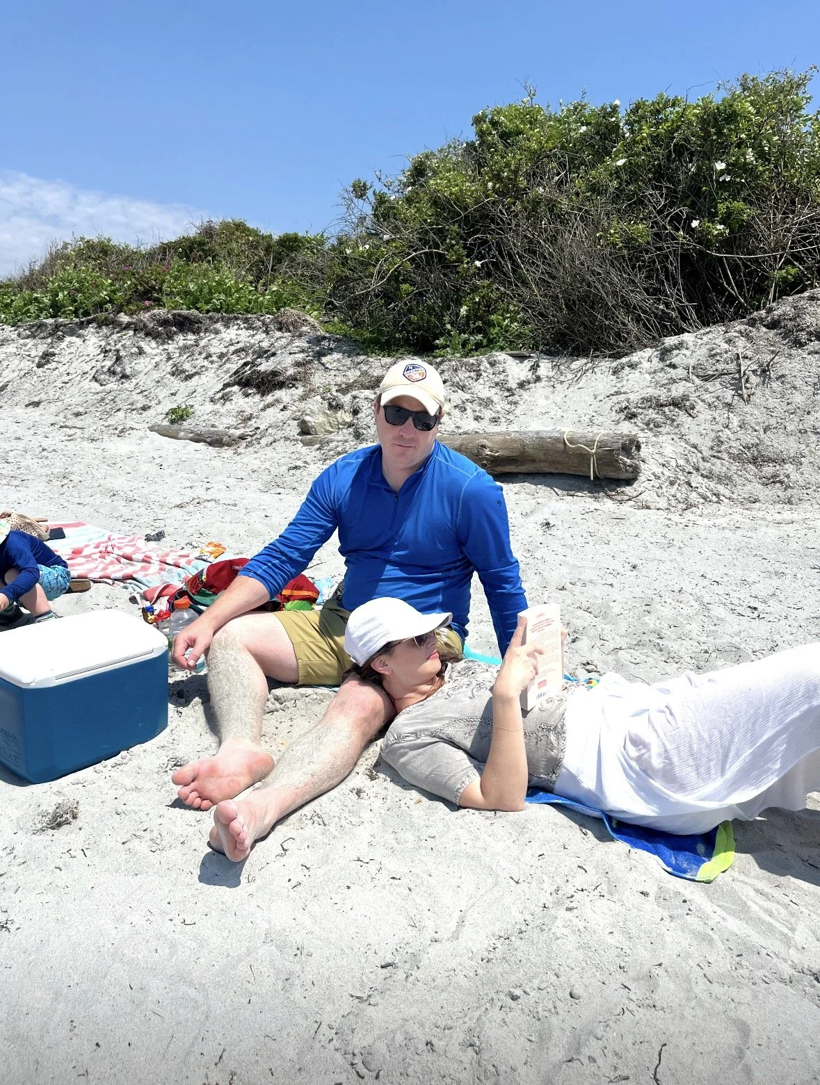 People relaxing on a sandy beach under a partly cloudy sky. One person is sitting with sunglasses and a hat, while another is lying down reading a book, both surrounded by beach gear and colorful towels.