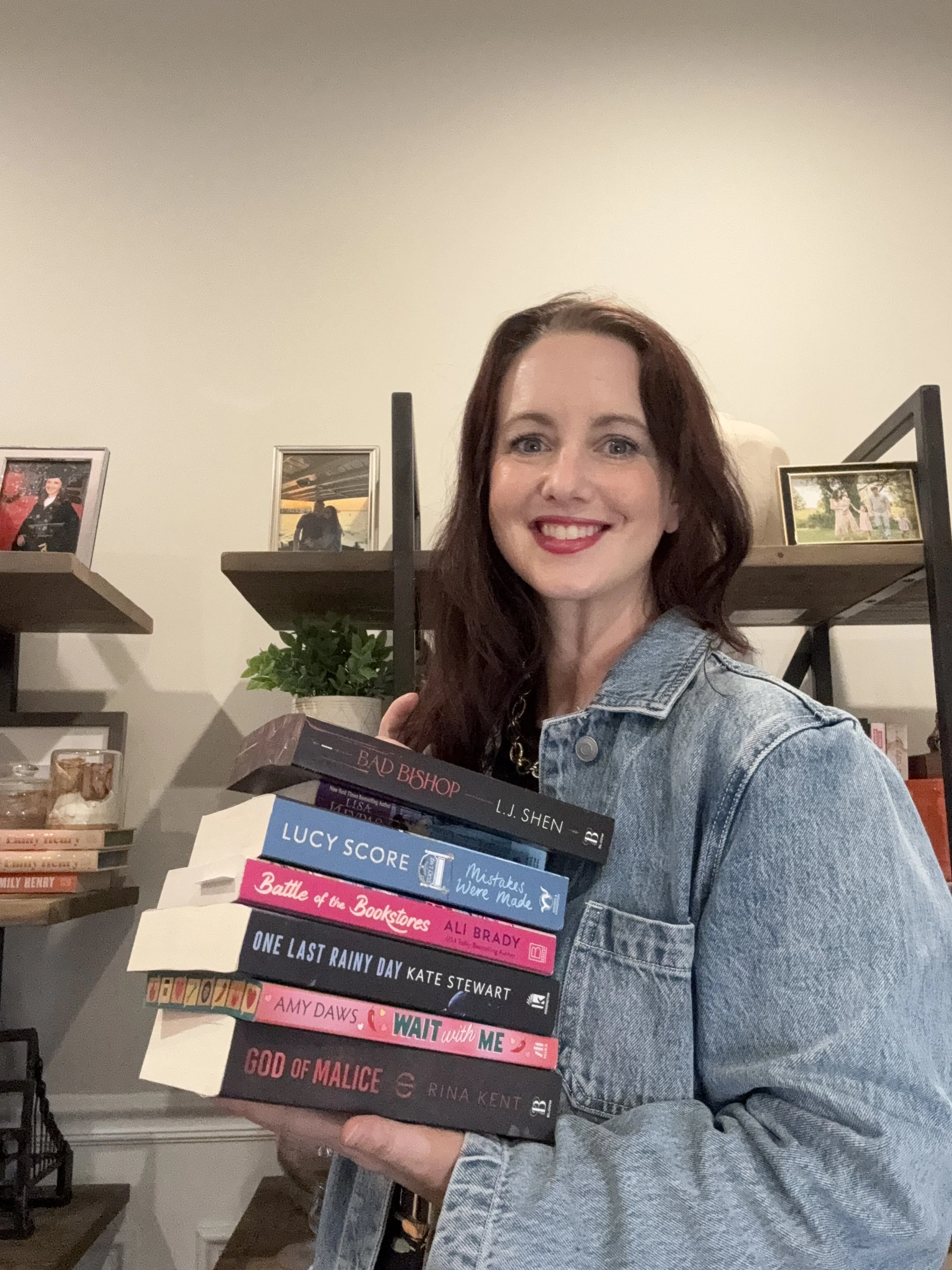 A woman with reddish-brown hair and a bright smile, wearing a denim jacket, holding a stack of books in a room with framed photos and shelves in the background.