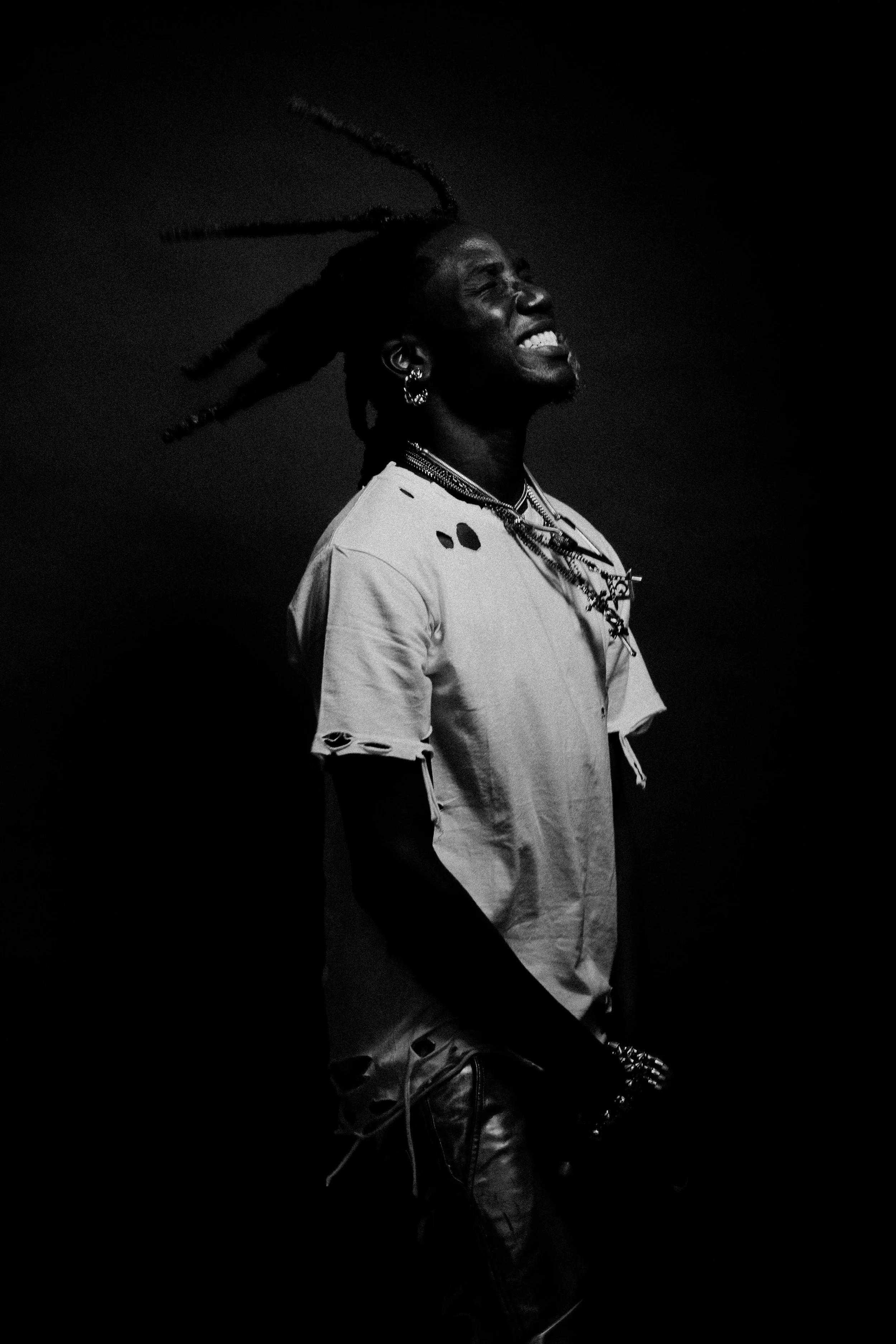 A black and white photo of a person with dreadlocks smiling and looking upwards. They are wearing multiple earrings, necklaces, a distressed t-shirt, and leather pants.