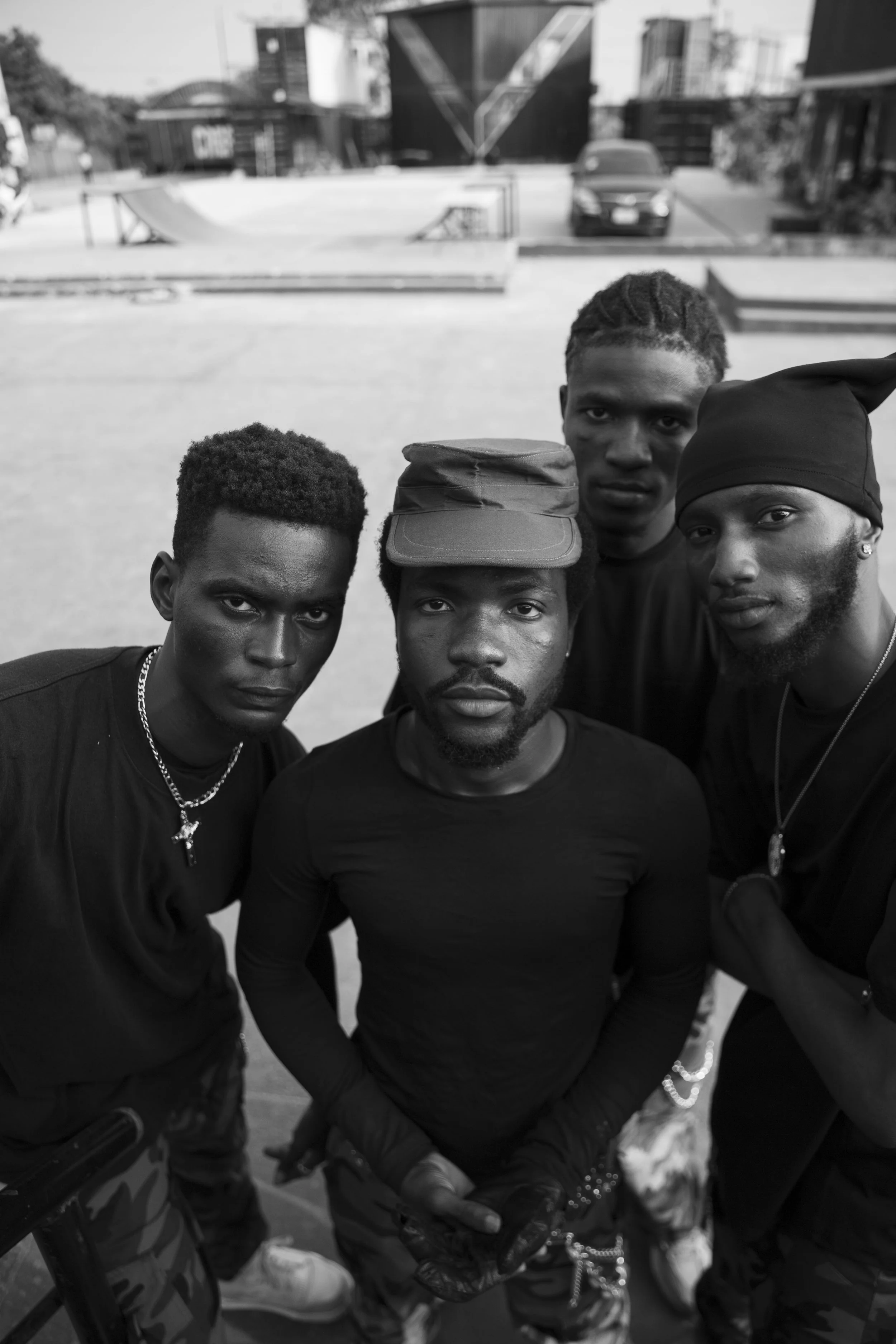 Four young men with dark skin looking into the camera in an outdoor urban area, with skate ramps and buildings in the background, all dressed in dark clothing and accessories.