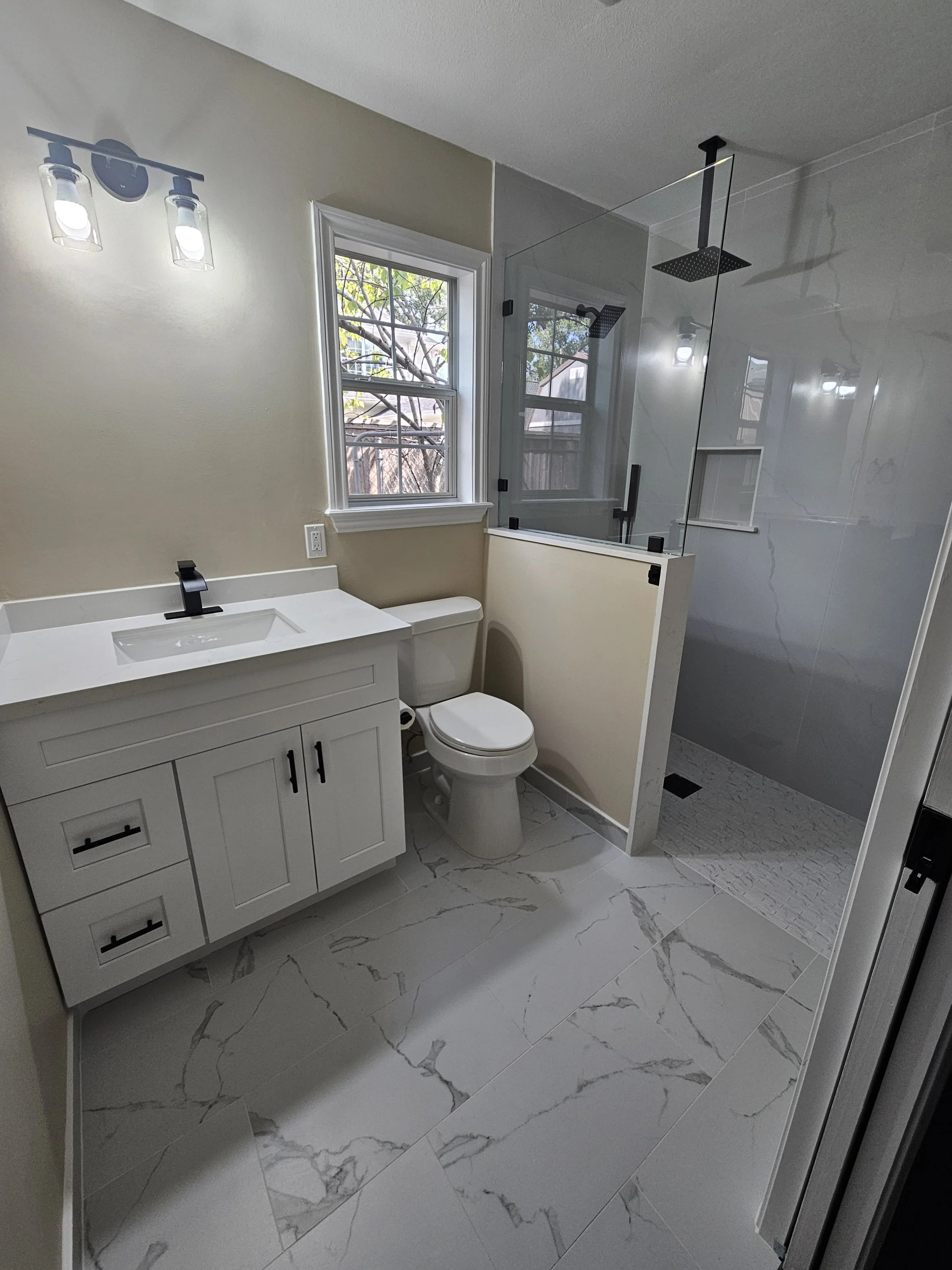 A modern bathroom with a white vanity, a toilet, a large window, and a glass enclosed shower with black fixtures. The floor is covered with white marble tiles with gray veining.