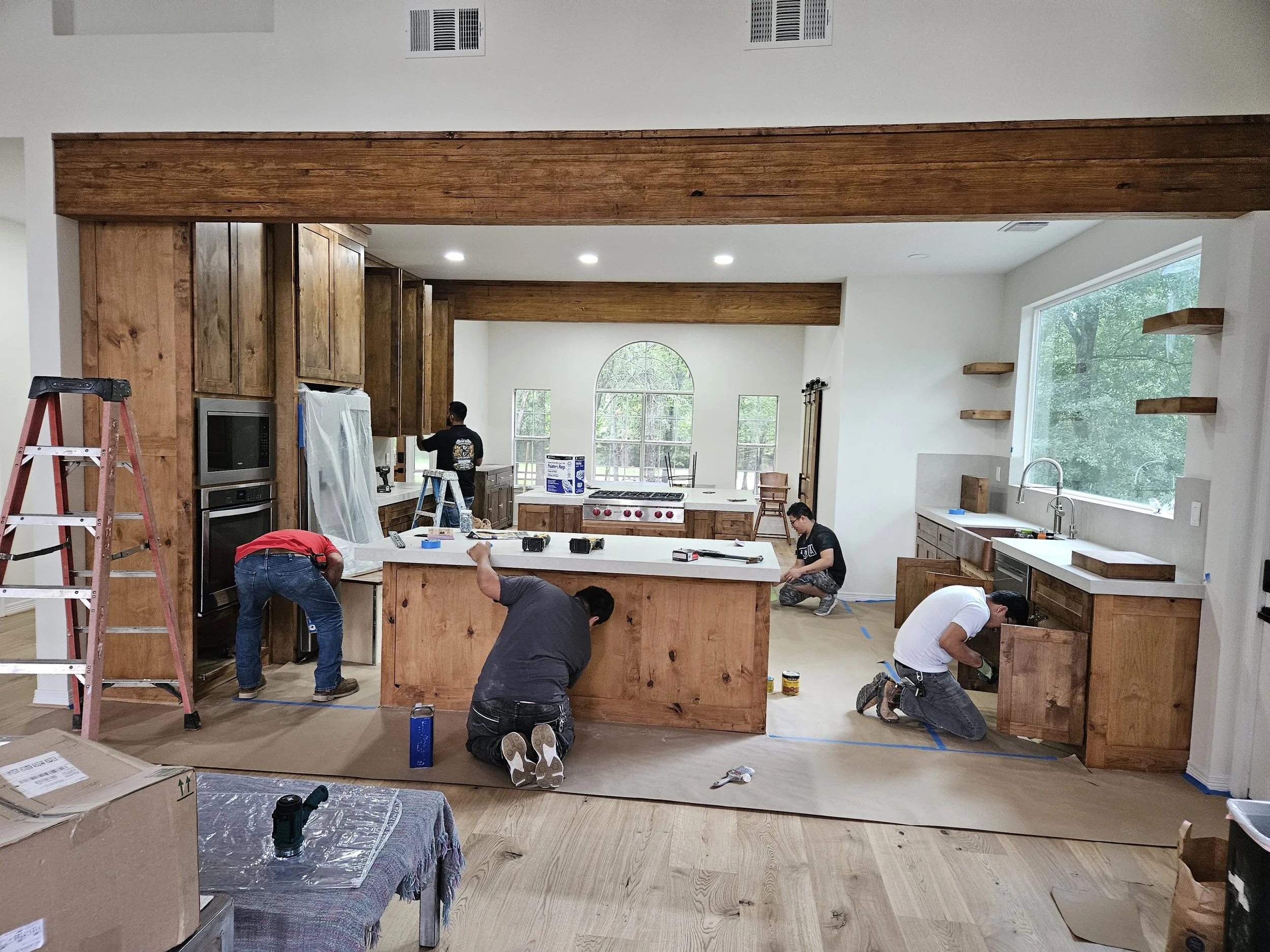 A kitchen under construction with workers installing cabinets, countertops, and appliances. There is a large island in the center, tools and materials scattered around, and large windows providing natural light.