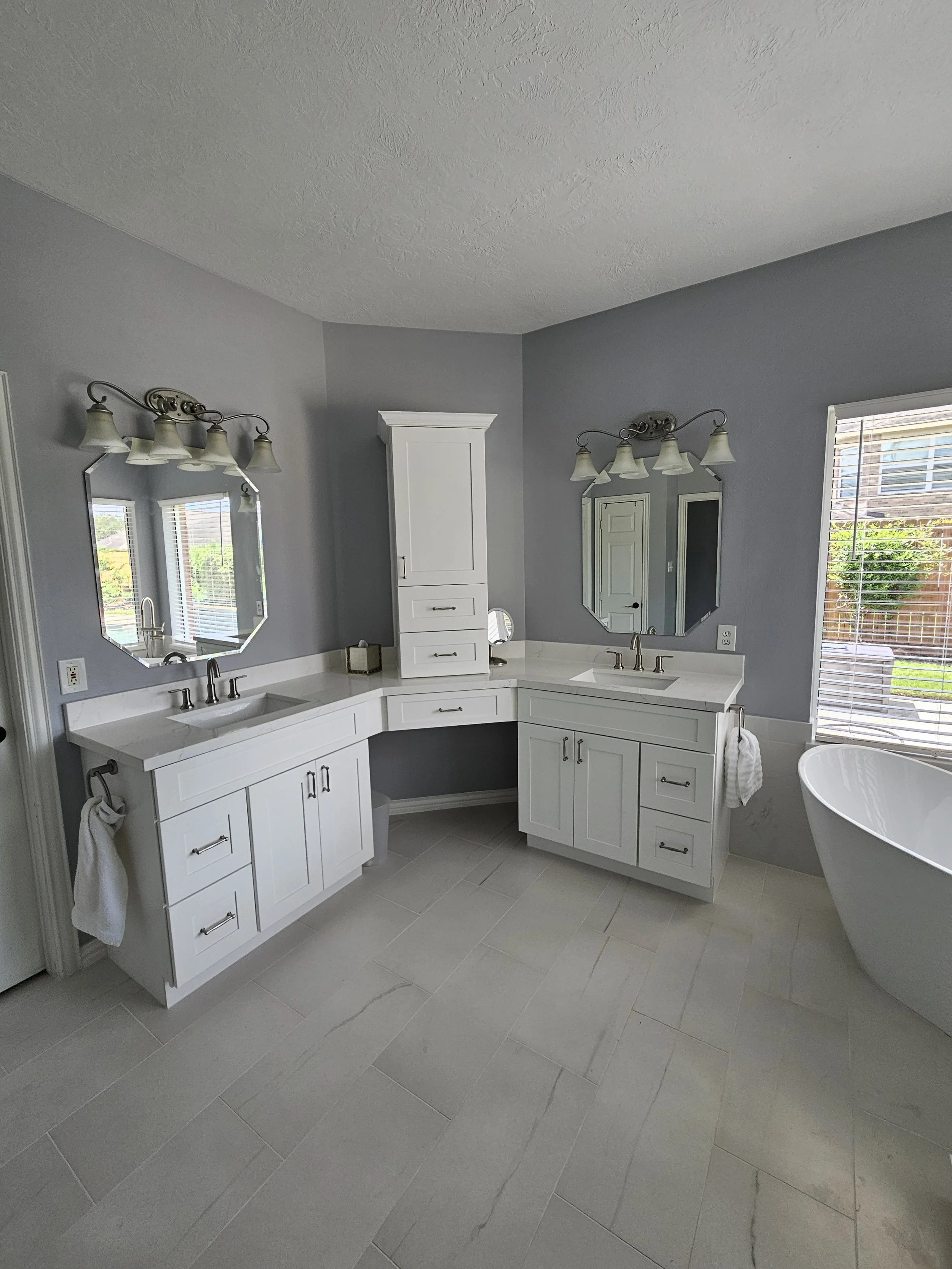 A modern bathroom with light gray walls and white cabinetry, featuring two mirrors above sinks, a white freestanding bathtub, and a window with wooden blinds, illuminated by two light fixtures with multiple lamps.