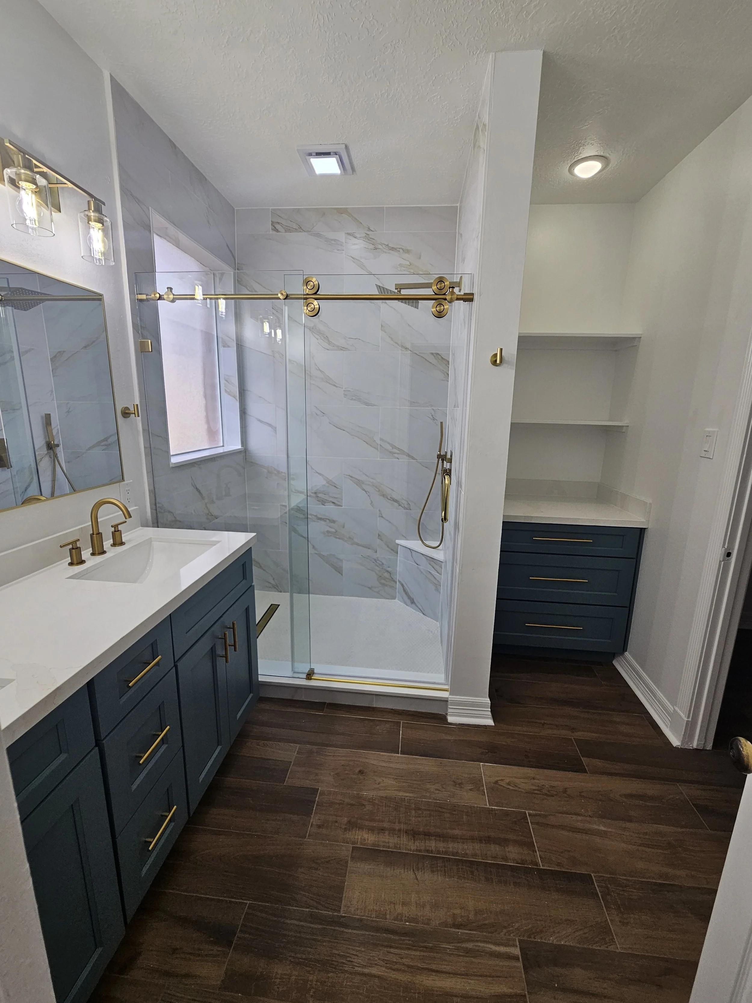 Modern bathroom with blue vanity, white marble countertop, brass fixtures, glass-enclosed shower with marble tiles, wooden floor, and built-in shelves.