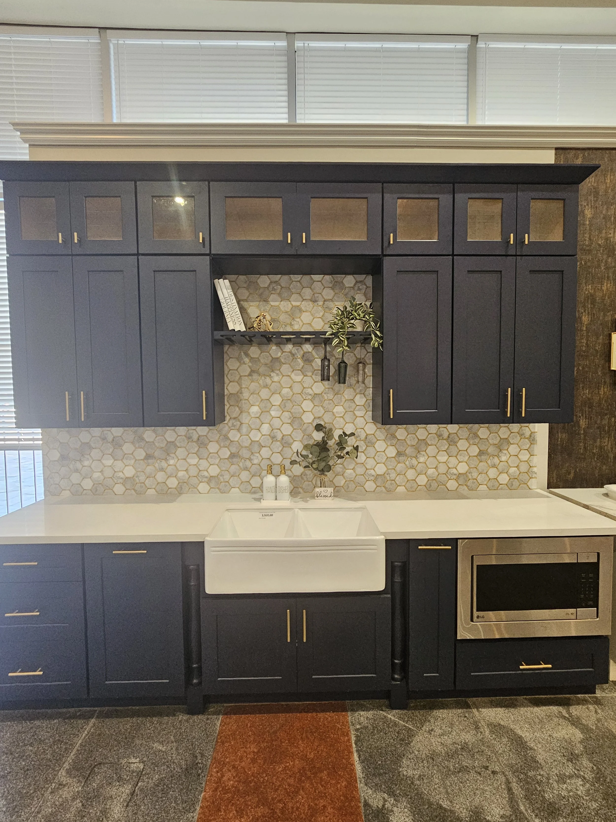Kitchen with blue cabinets, white farmhouse sink, honeycomb tile backsplash, and black legs on the cabinets. Decor includes a small potted plant, books, and decorative items. The floor is tiled with dark and rust-colored stones.