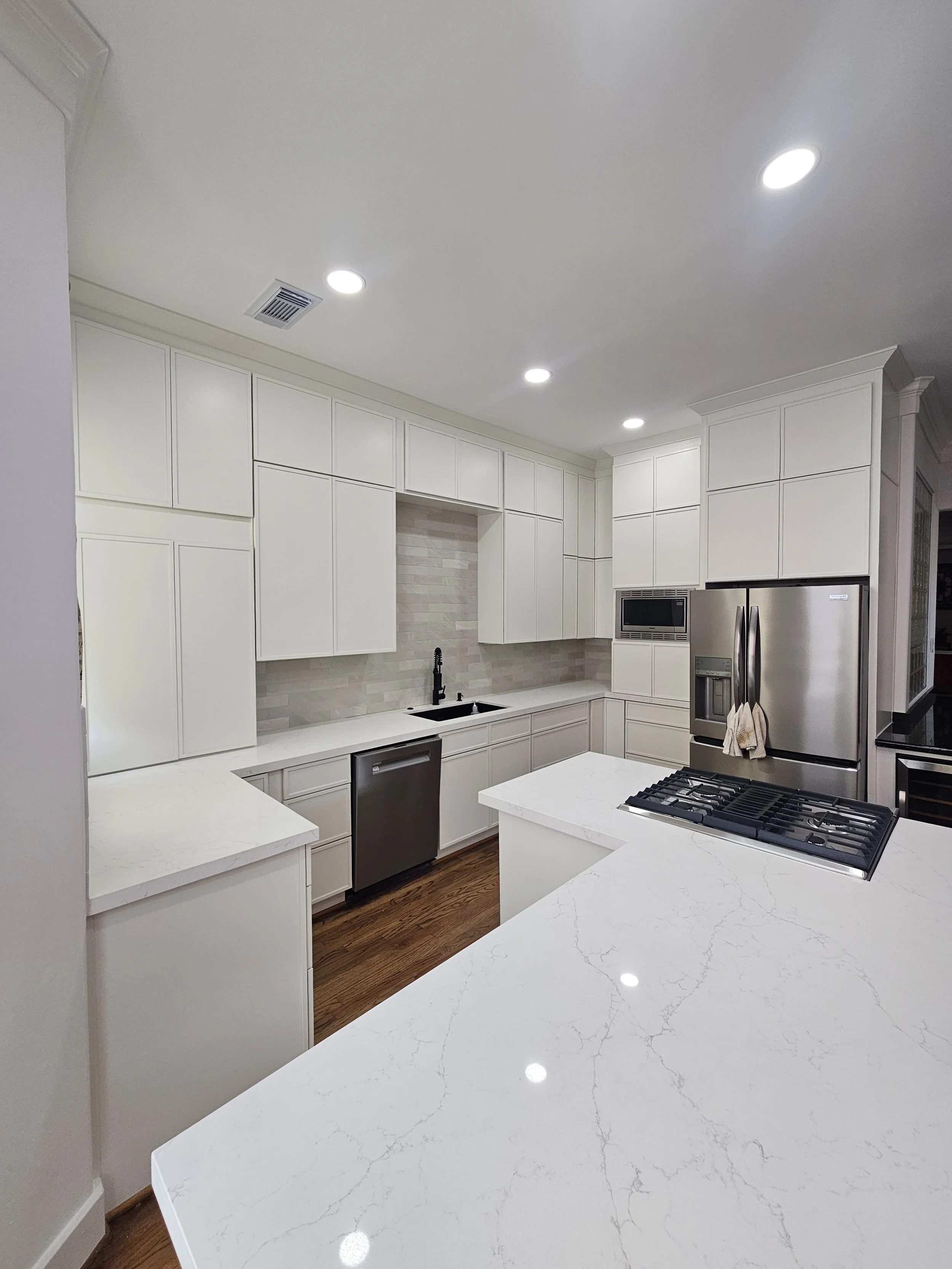 Modern kitchen remodeling with white cabinets, marble countertop island, stainless steel appliances, and hardwood flooring, illuminated by ceiling lights.