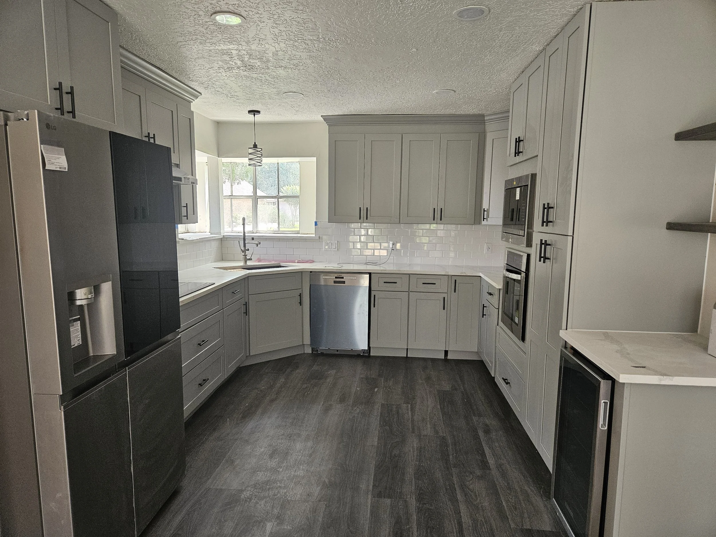 Modern kitchen with gray cabinets, white subway tile backsplash, stainless steel appliances, including a double oven and refrigerator, and a large window above the sink letting in natural light. Dark wood flooring completes the space.
