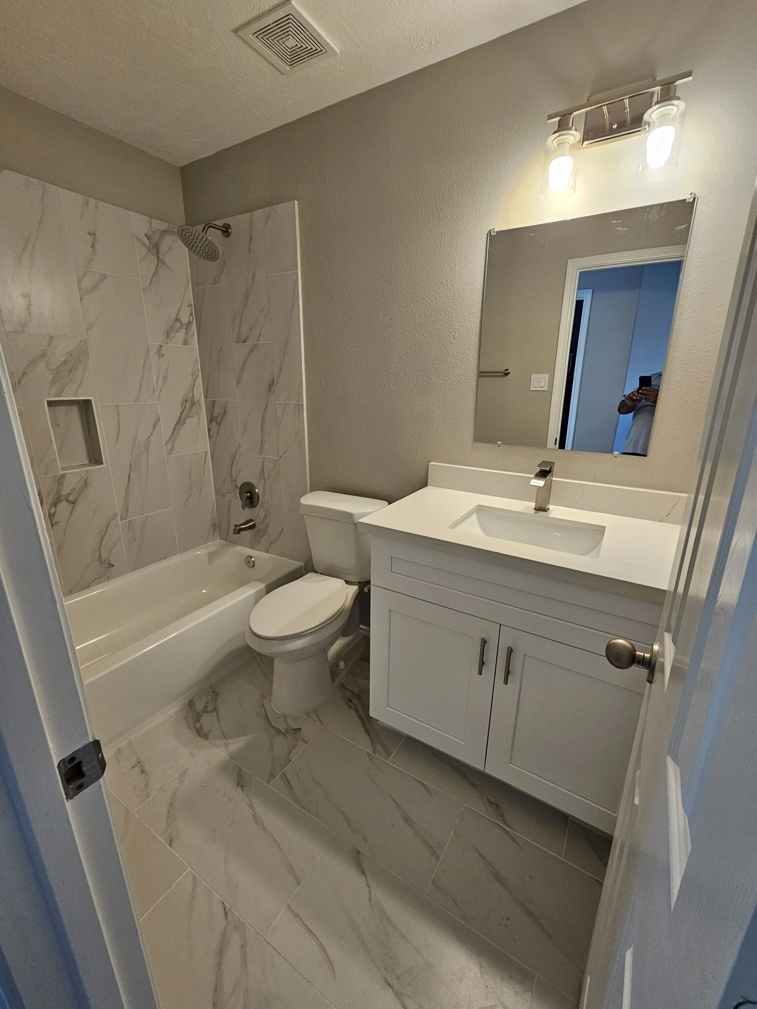 A bathroom with a bathtub and shower on the left, a toilet in the middle, and a white vanity with a sink and mirror on the right. The bathroom has marble-like tiled floors and walls, a small built-in shower shelf, and a light fixture above the mirror