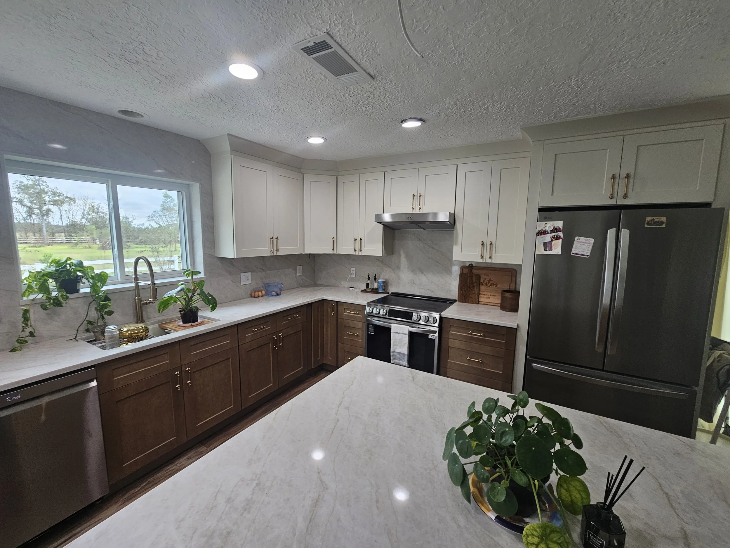 Modern kitchen with white upper cabinets, brown lower cabinets, marble countertops, stainless steel stove and range hood, large window overlooking a green outdoor area, and several potted plants on the counter.