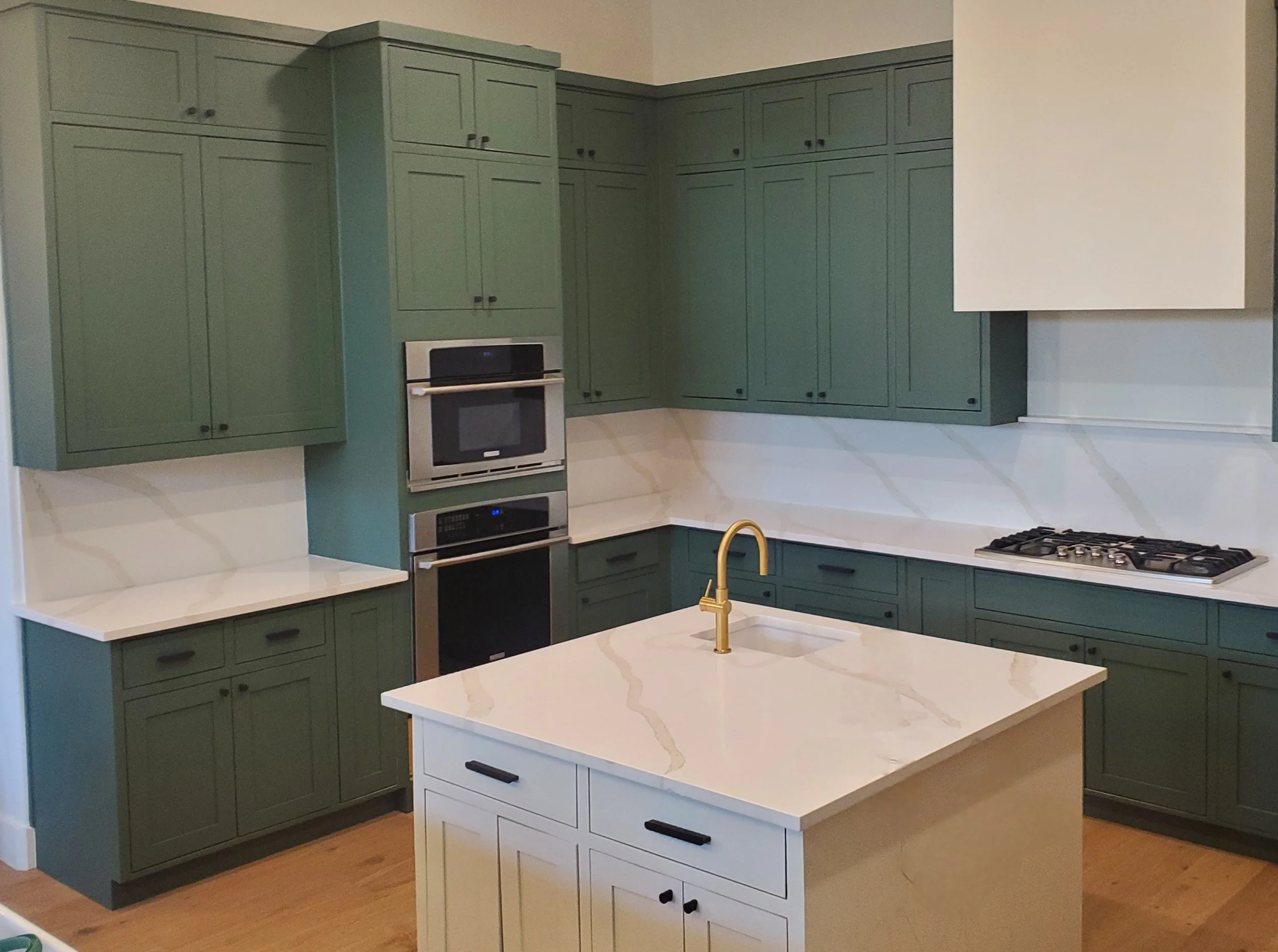 Kitchen with green cabinets and white countertops, built-in oven, microwave, and stovetop, island with white marble-style surface, sink with yellow faucet.