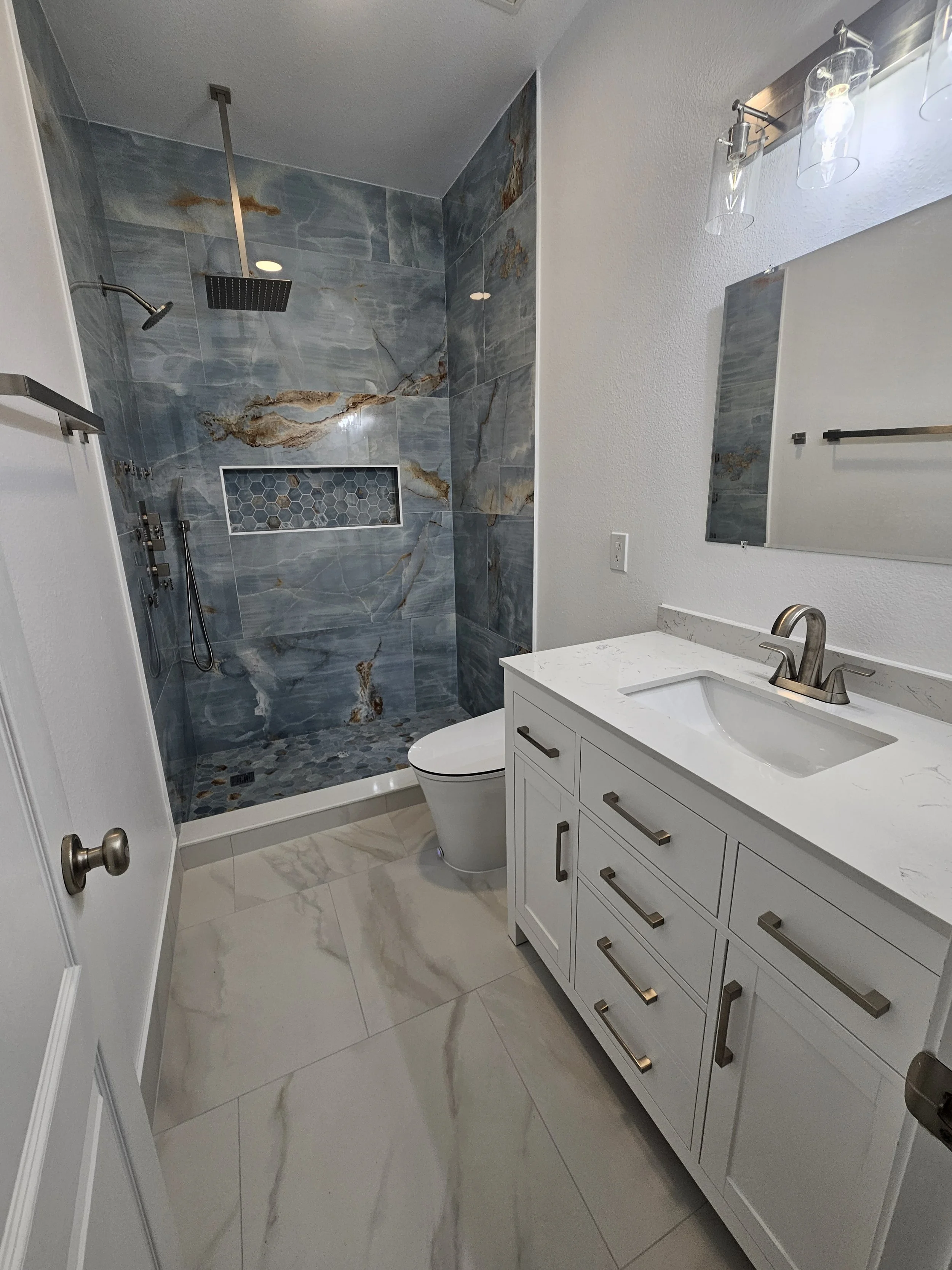 Modern bathroom with white vanity, marble countertop, large mirror, and walk-in shower with blue-gray marble tiles, hexagonal tile accent, and rainfall showerhead.