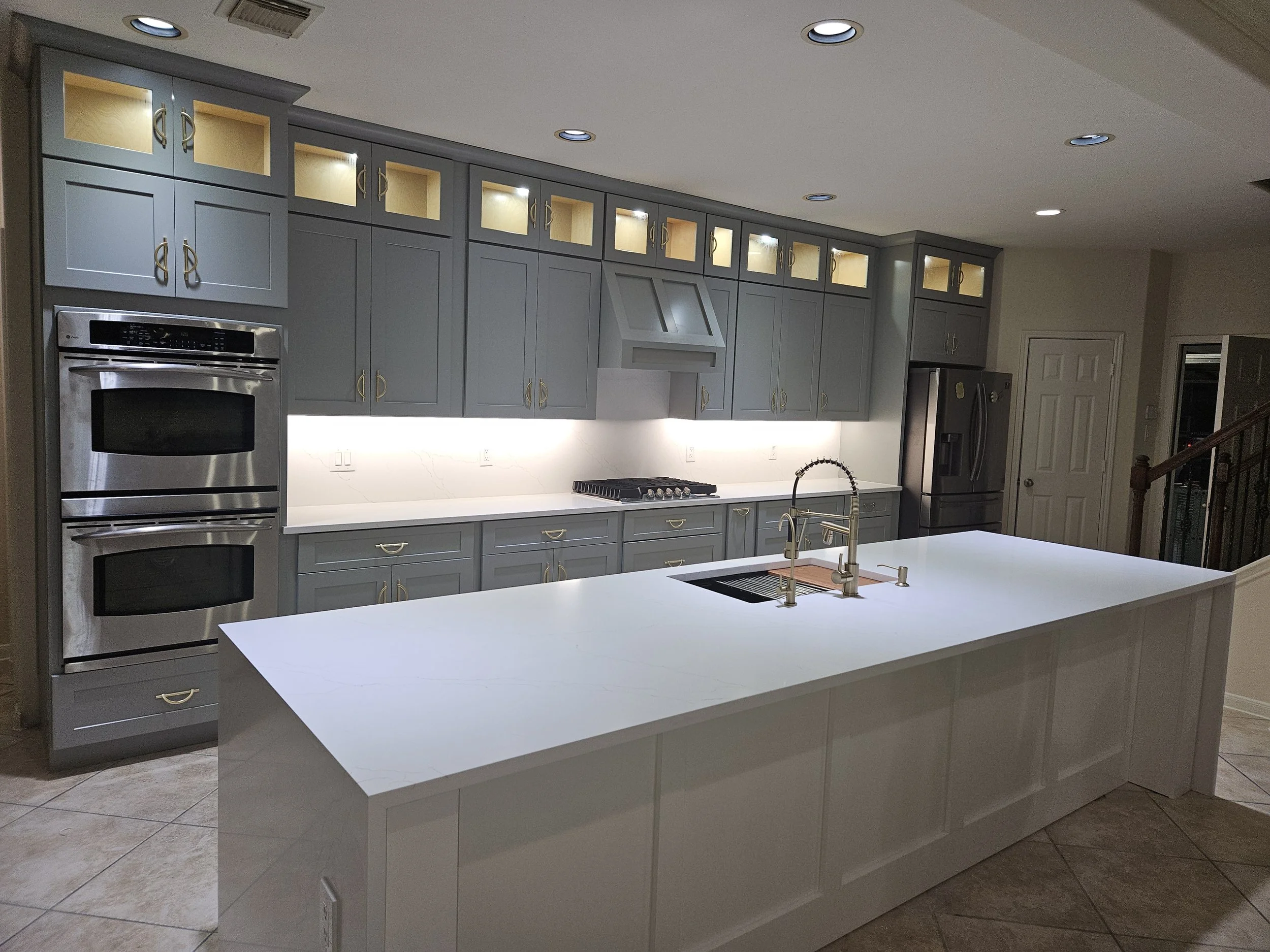 A modern kitchen with gray cabinets, a white countertop island, stainless steel appliances, and recessed ceiling lighting.