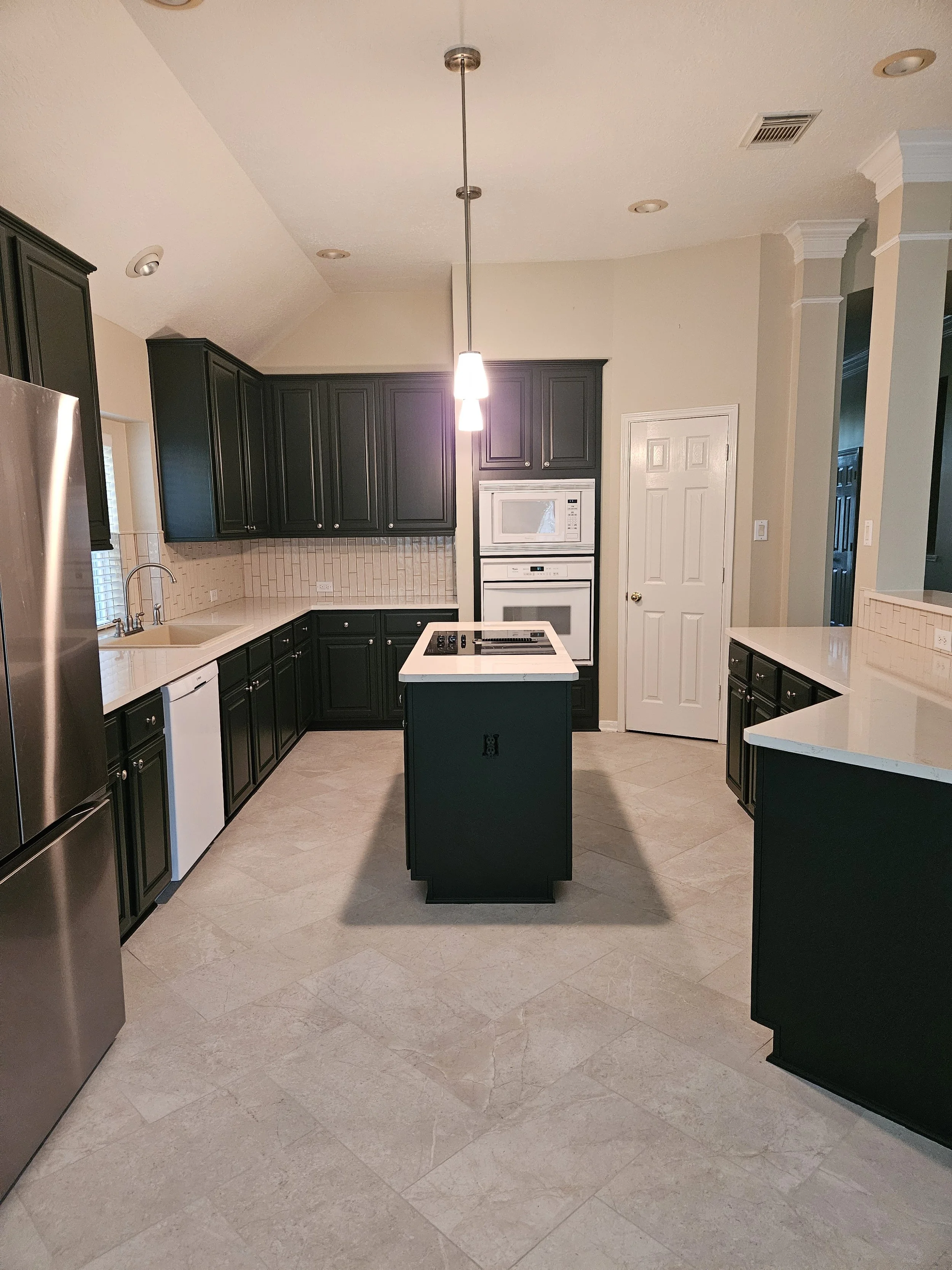 A kitchen with black cabinets, beige countertops, white appliances, and beige tile flooring. A white door and a mosaic tile backsplash are visible on the wall. A small island with a stovetop is in the center, and a hanging light fixture is above.
