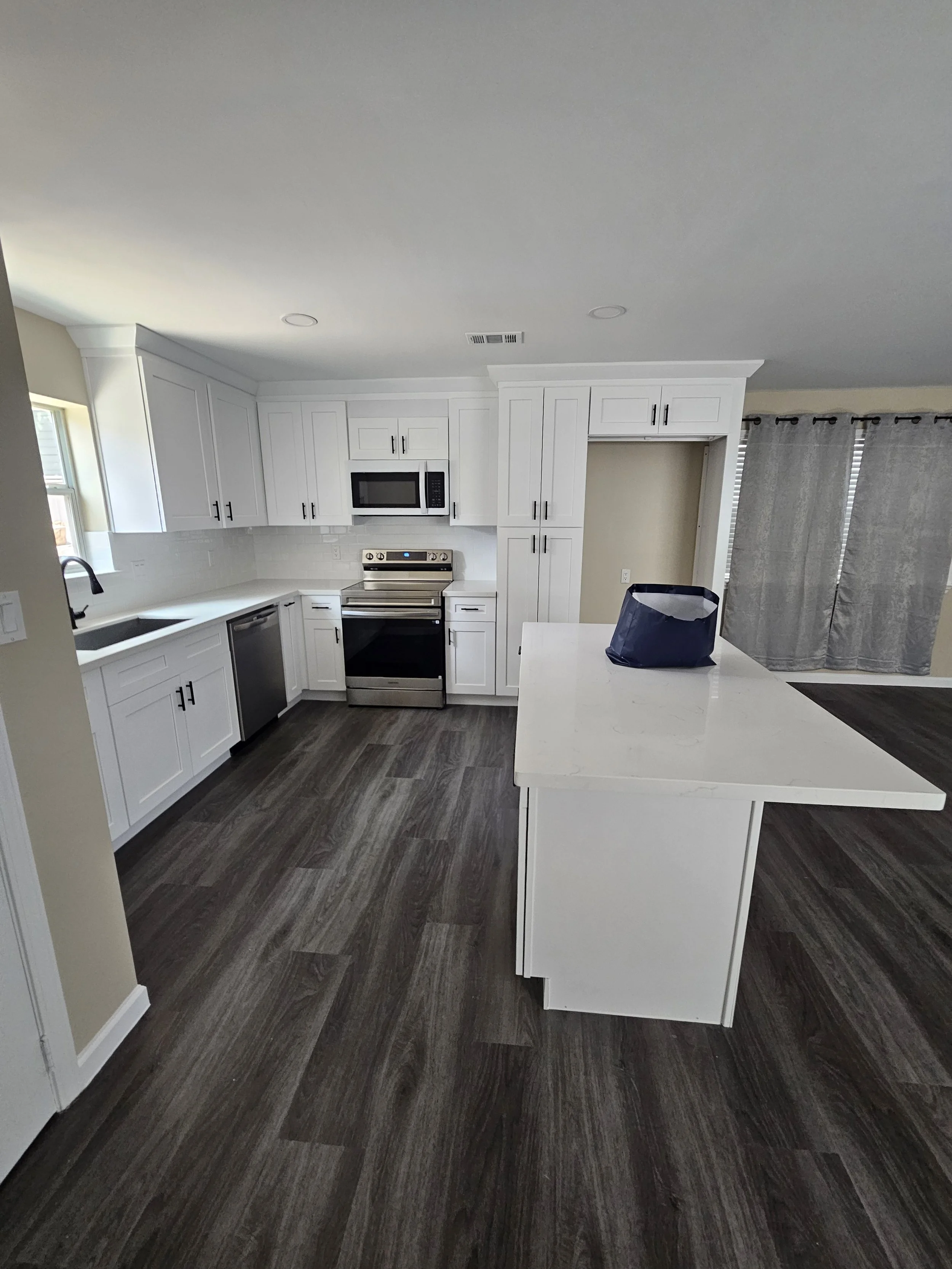 Modern, white kitchen with dark wood flooring, stainless steel appliances, white cabinetry, and a kitchen island with a black bag on it.