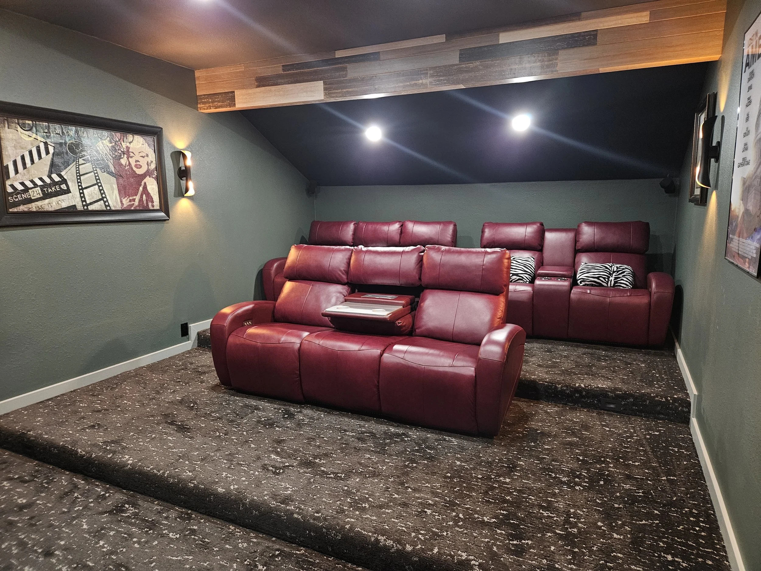 Construction A theater room with green walls, a black ceiling, a wooden accent above, and a maroon leather sofa and recliner with pillows. There are framed pictures and wall sconces on the walls, and the floor has a patterned carpet.