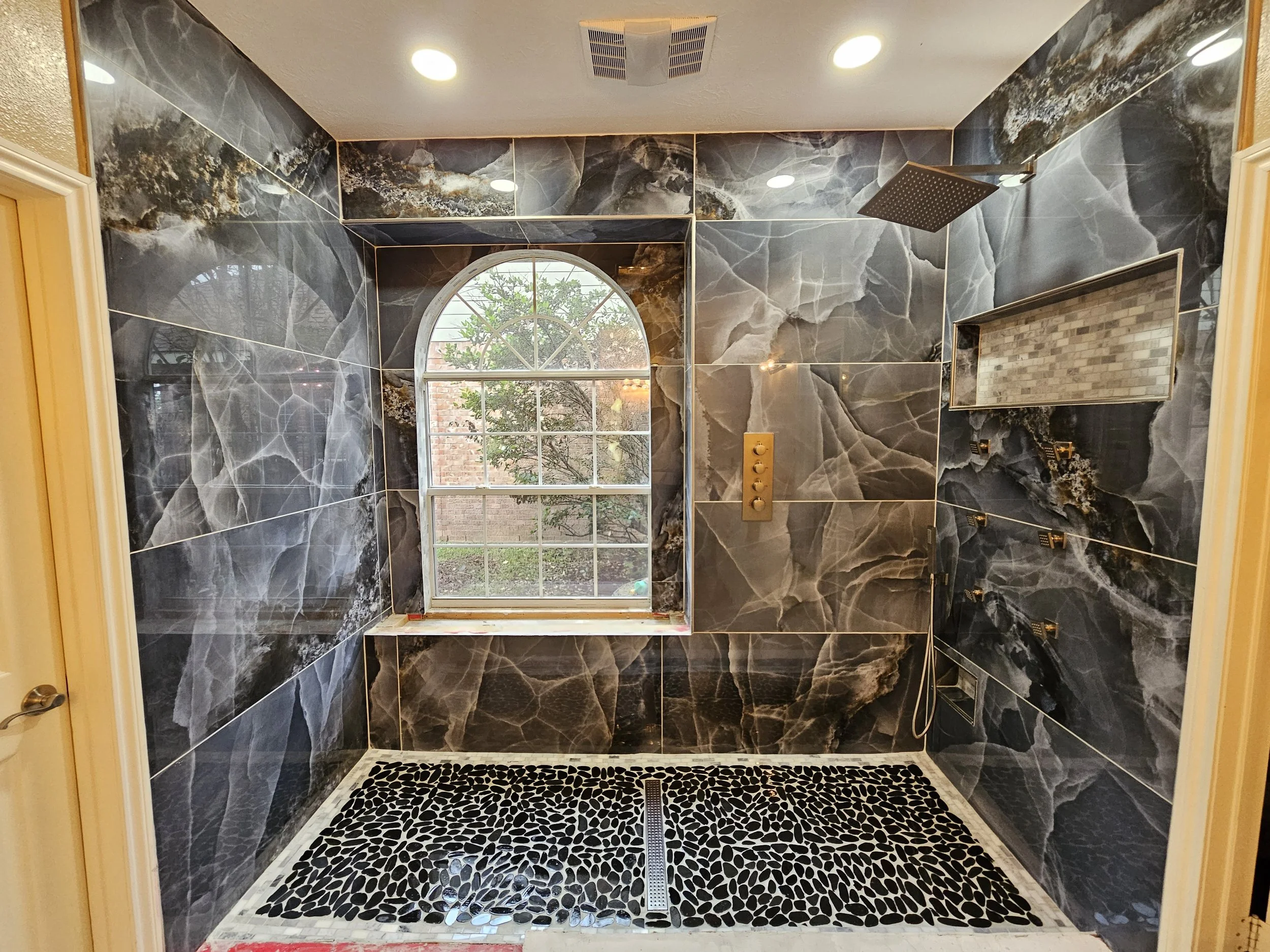 A modern shower with dark marble tiled walls, a large rainfall showerhead, a built-in niche with a pebble floor, and a window showing a backyard with trees and a brick wall.