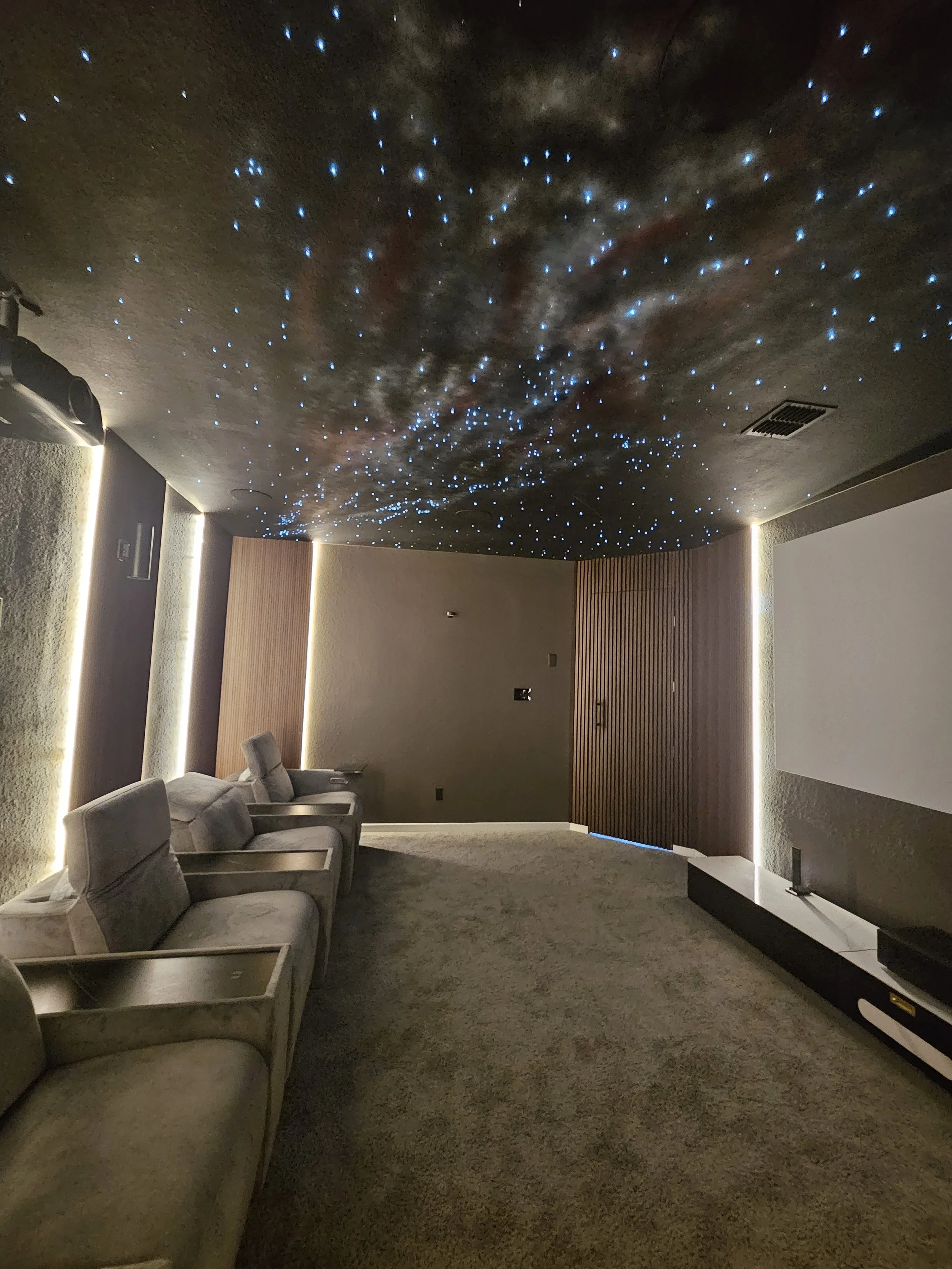 A room with a starry sky ceiling featuring blue LED lights, beige sofas on the left, wooden wall panels at the back, and gray carpeted floor.