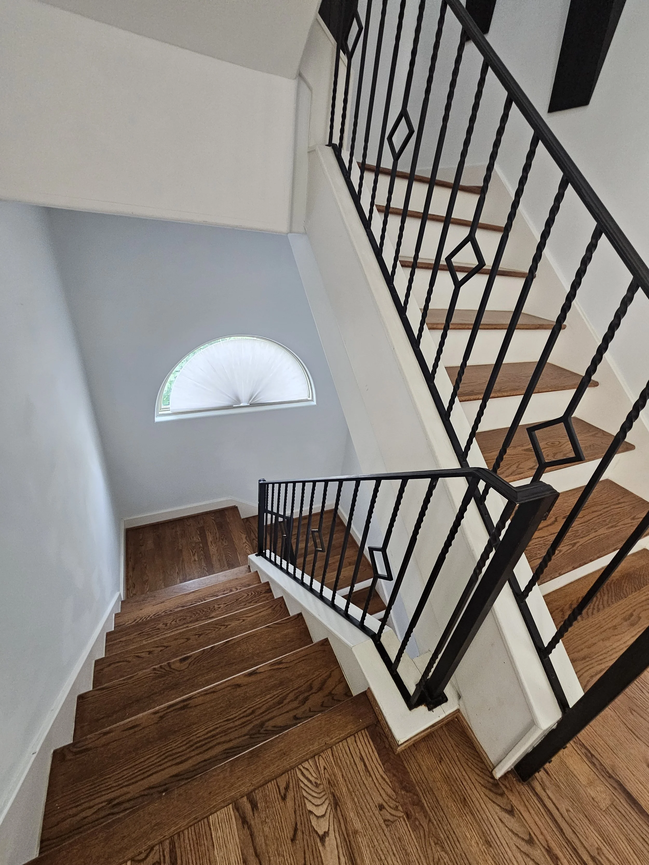 Interior view of a staircase with wooden steps, black iron railings, white walls, and a semi-circular window on the wall letting in natural light.