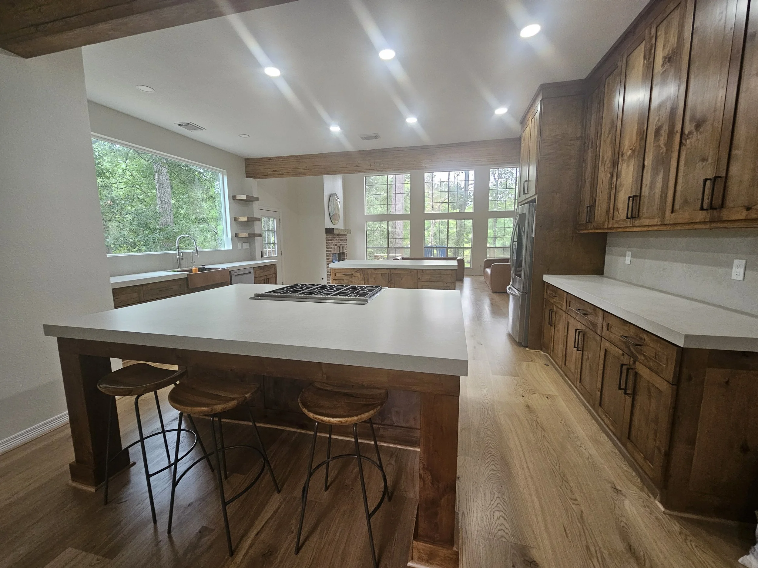 Home Renovation with Open kitchen with white island, wooden cabinets, and large windows showing greenery outside, with three wooden bar stools at the island.