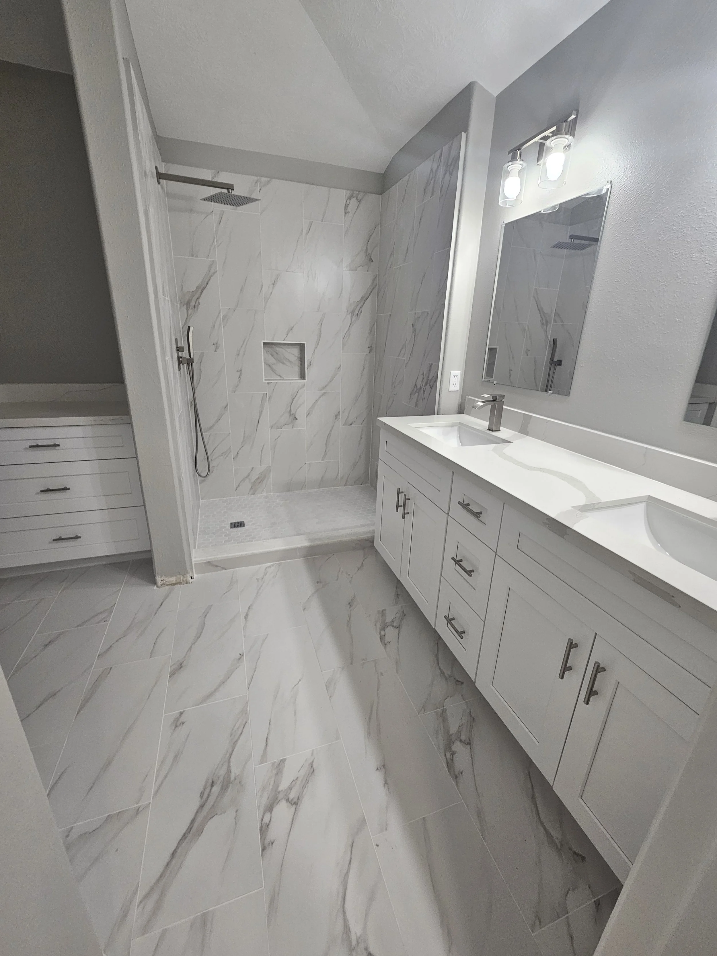 Bathroom with shower area featuring gray and white marble tiles, a large mirror above a white vanity with dual sinks, and modern light fixtures.
