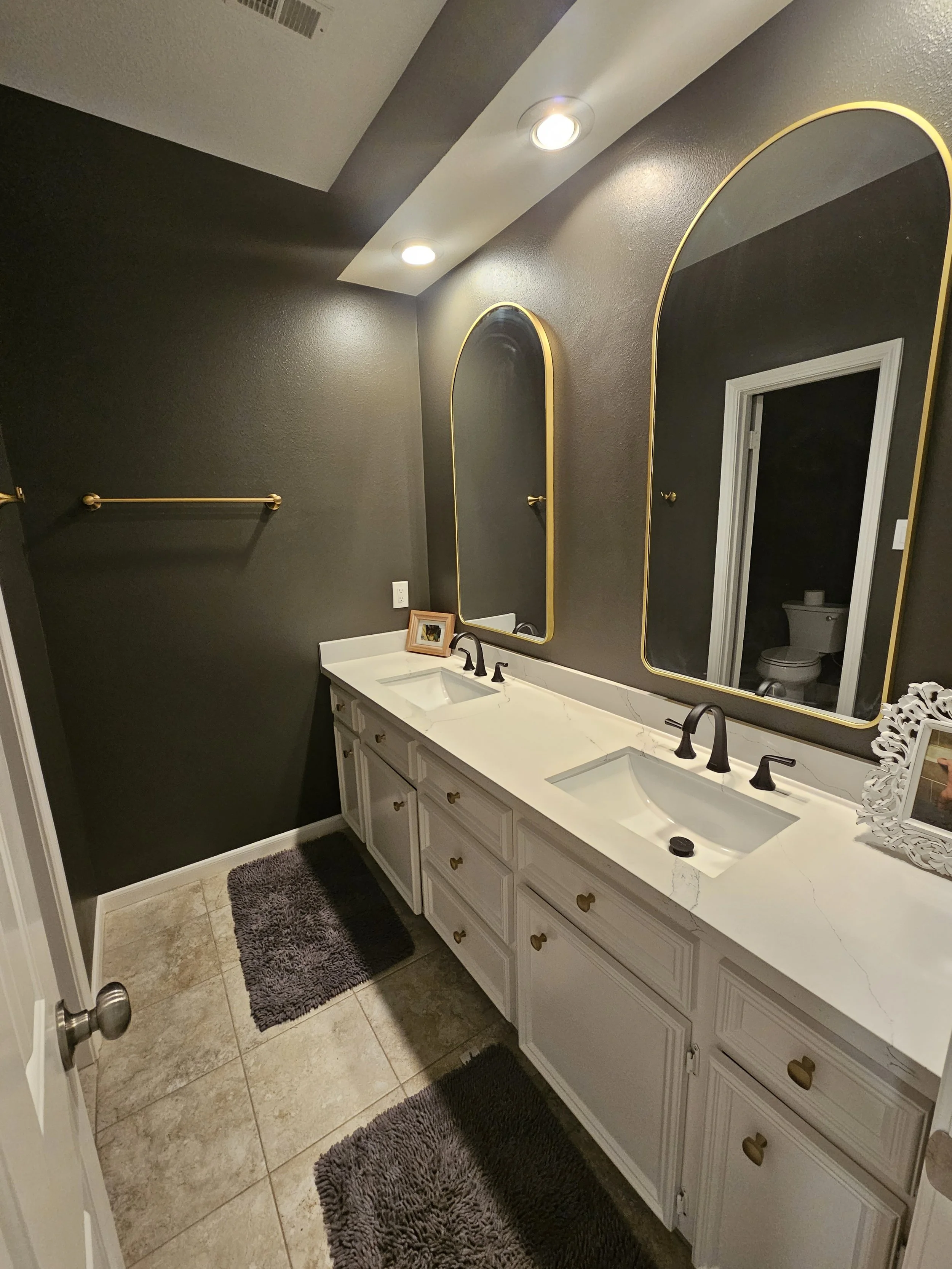 Bathroom with dark gray walls, white vanity with two sinks, large mirror with gold trim, and two ceiling lights. There are two black towels on a gold towel bar, a small picture frame on the vanity, a decorative white picture frame, and a gray rug on 